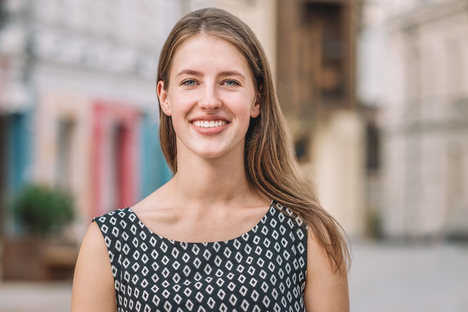Bright and cheerful portrait of a young woman standing on a city street, smiling at the camera.