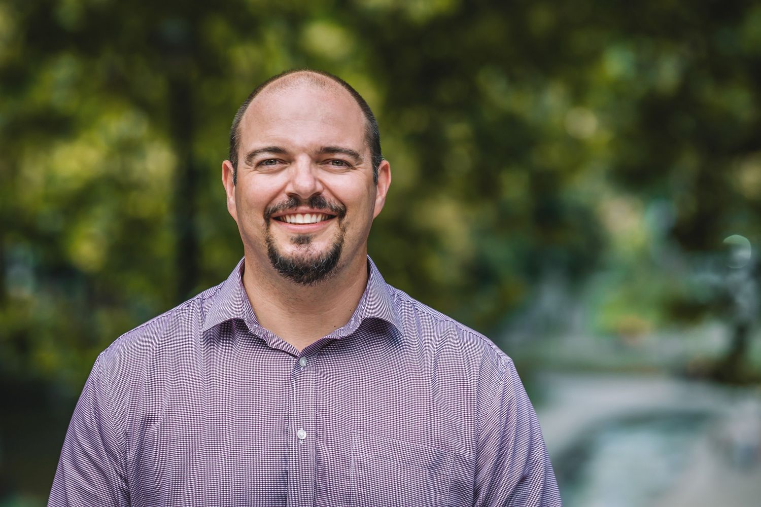 Bald man with a friendly smile, photographed outdoors with greenery in the background.