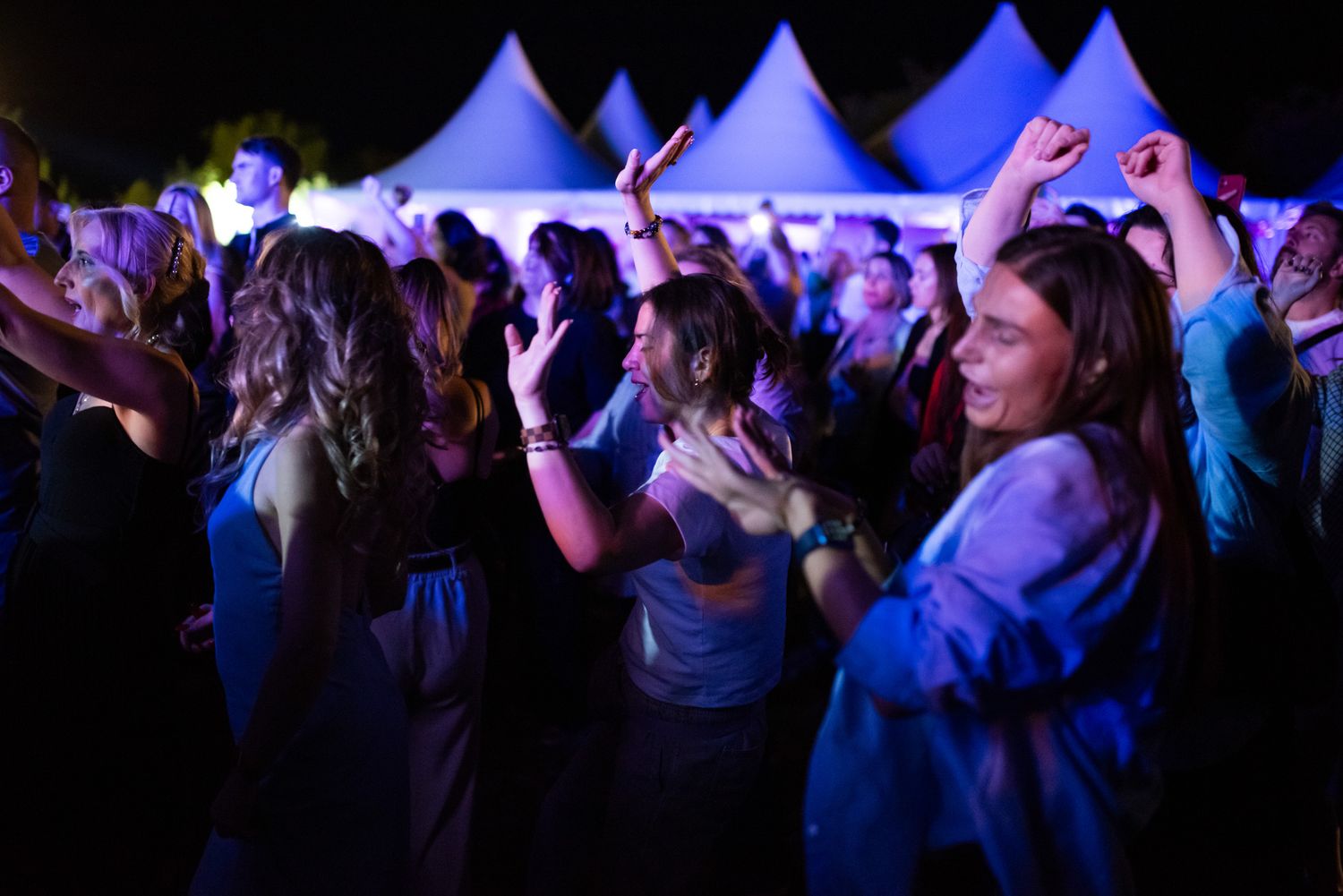 A crowd enjoying a night concert or festival, people dancing and raising their hands under white event tents.