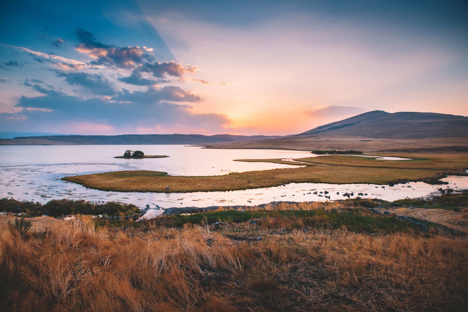 Dramatic sunset over a calm lake and grassy shoreline, with distant mountains in the background.