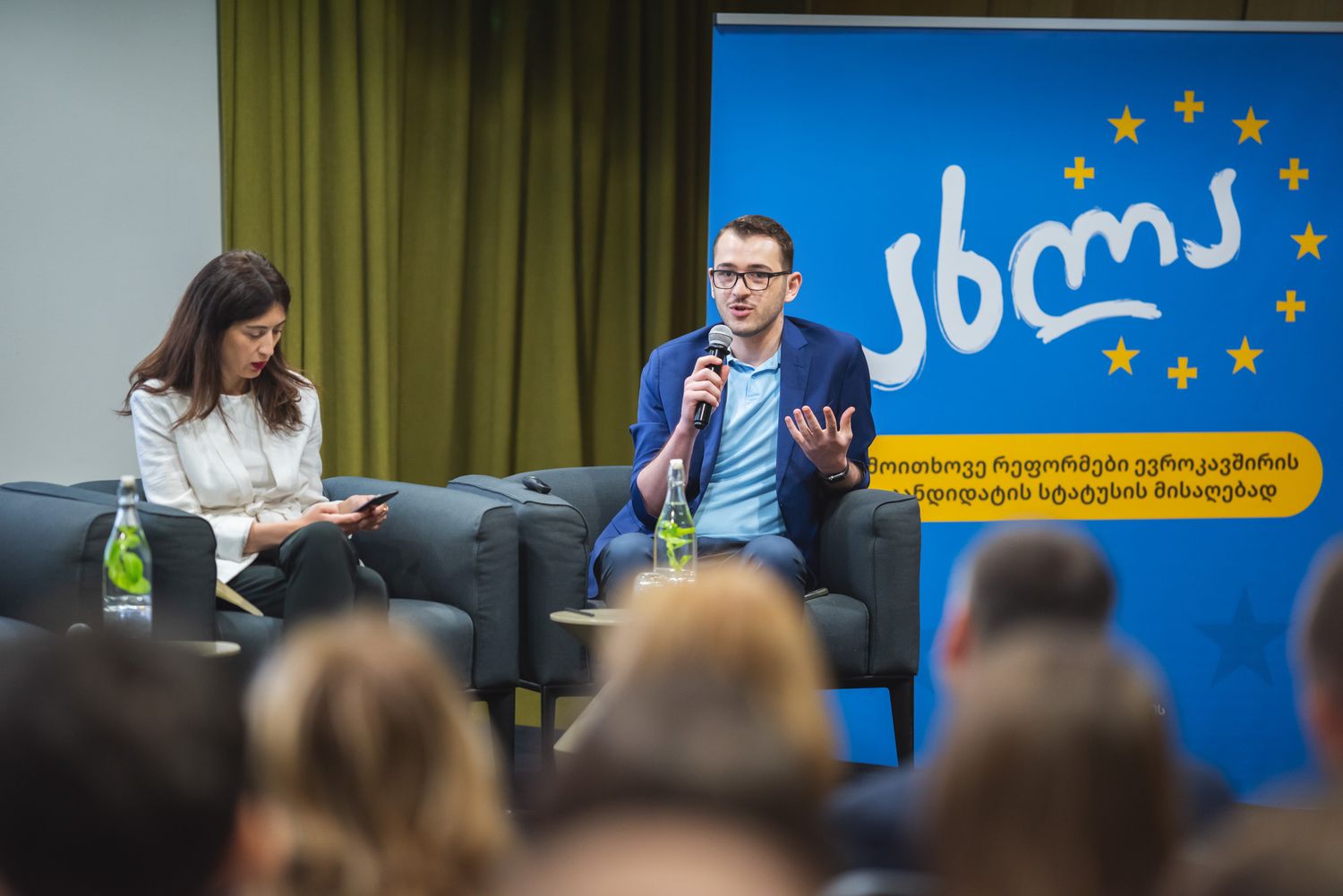 Panelists participating in a live event or talk show, with a branded blue and yellow backdrop.