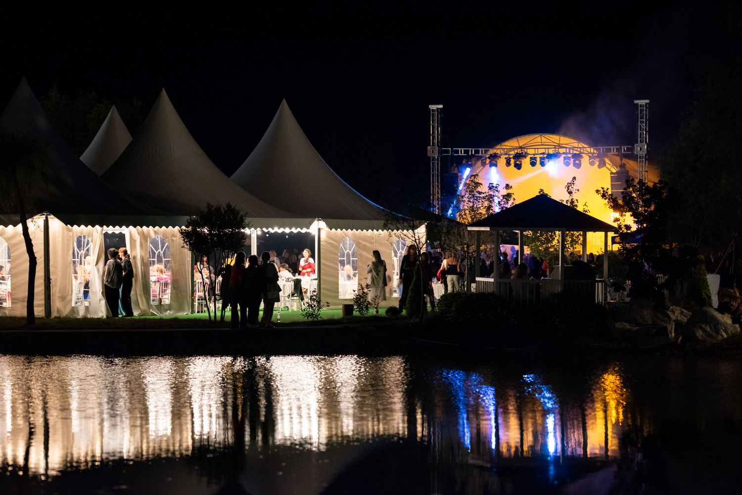 Scenic night view of illuminated event tents reflected on a calm water surface.