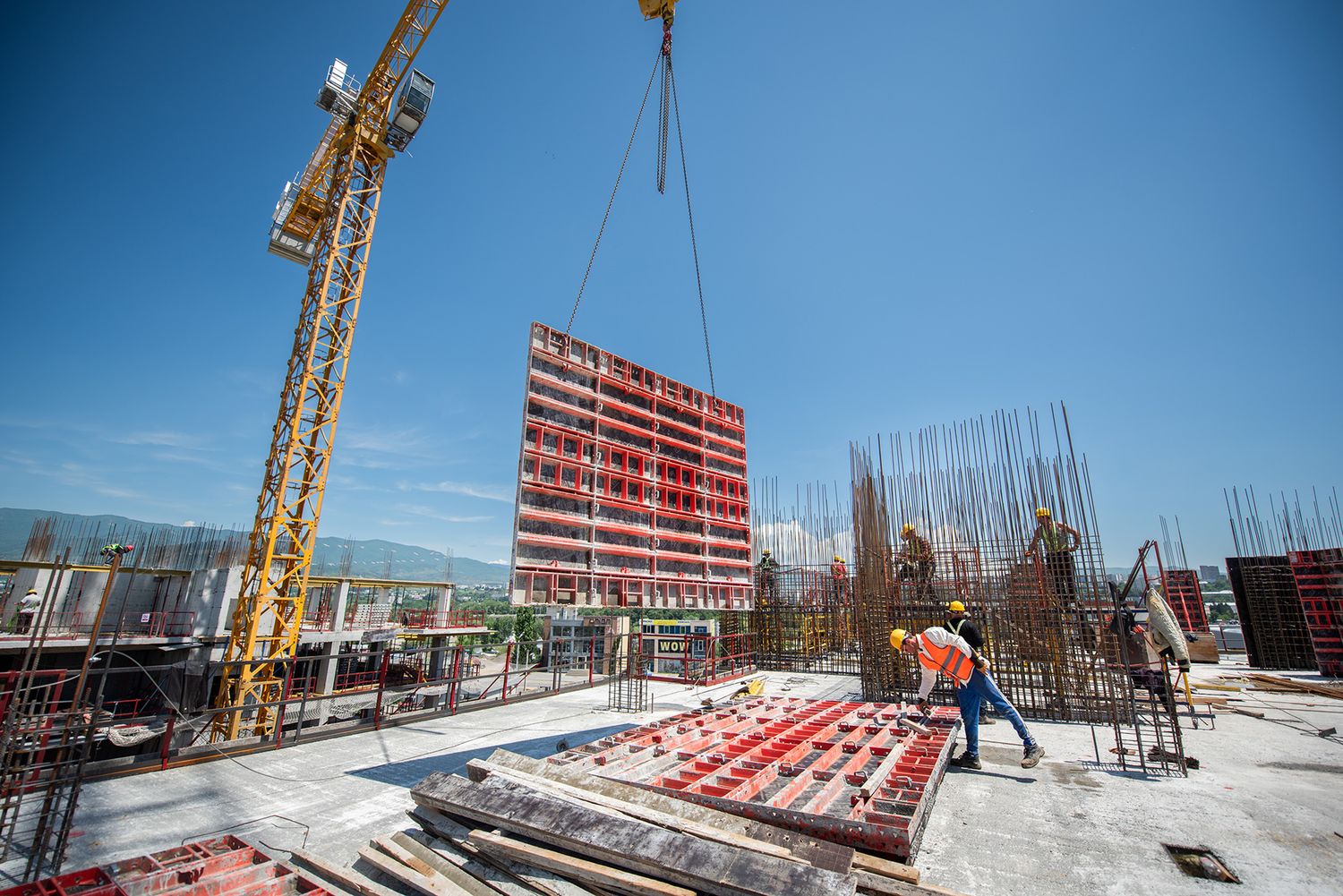 Construction crane lifting red metal formwork panels at a large-scale concrete site.
