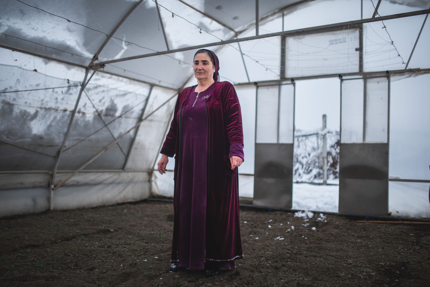 A priest or monk standing solemnly inside a greenhouse, wearing traditional maroon robes.