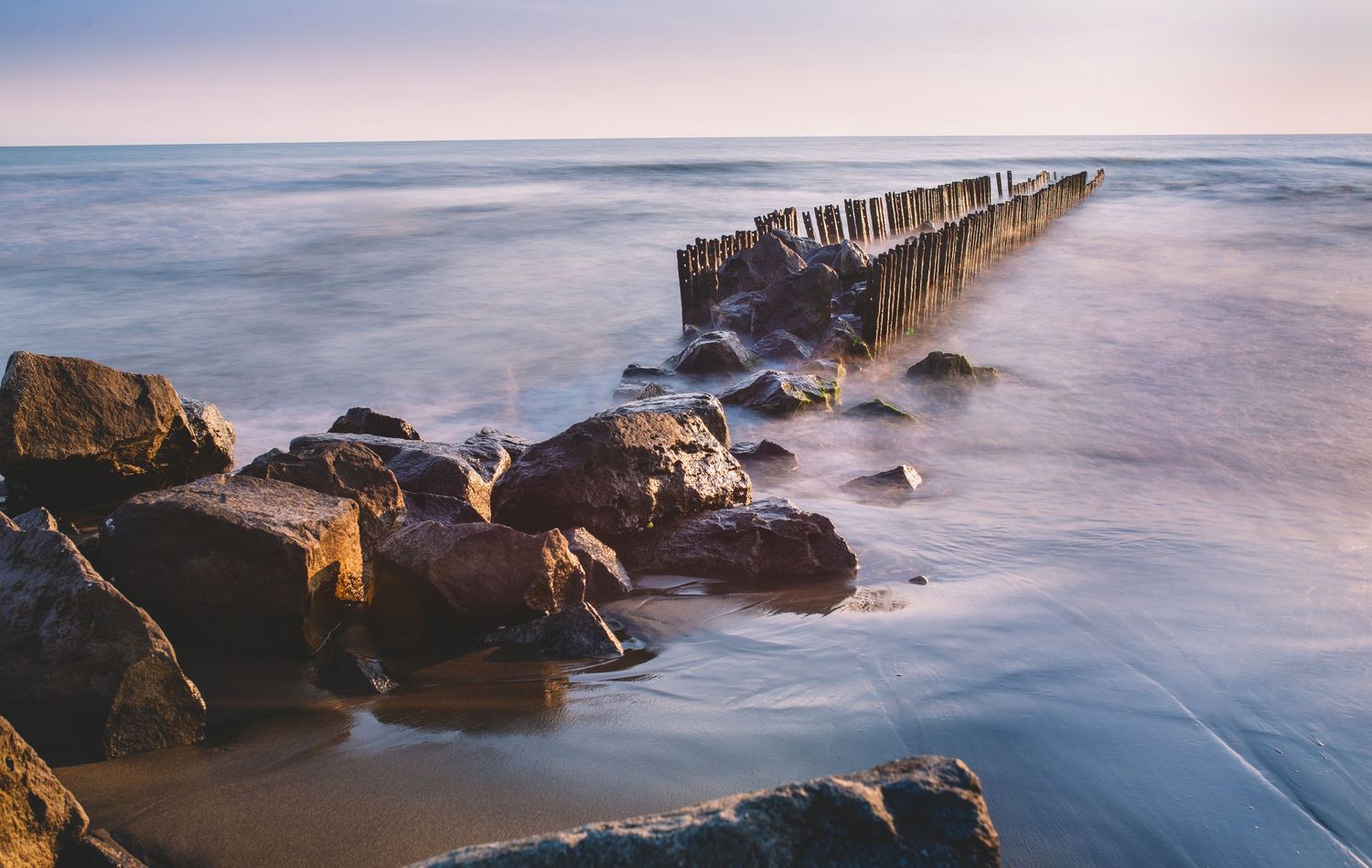 Rocky shoreline at sunset with gentle waves and pastel sky hues — serene seascape scene.