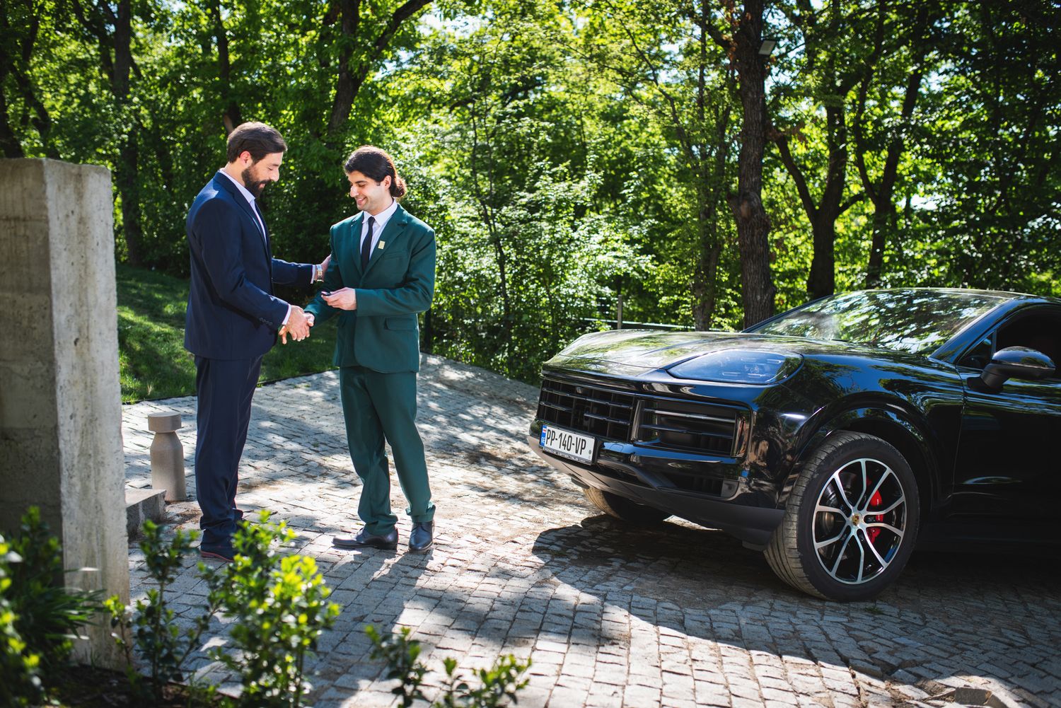 Two men in suits gesturing beside a luxury car — busia ness meeting or negotiation scene outdoors.