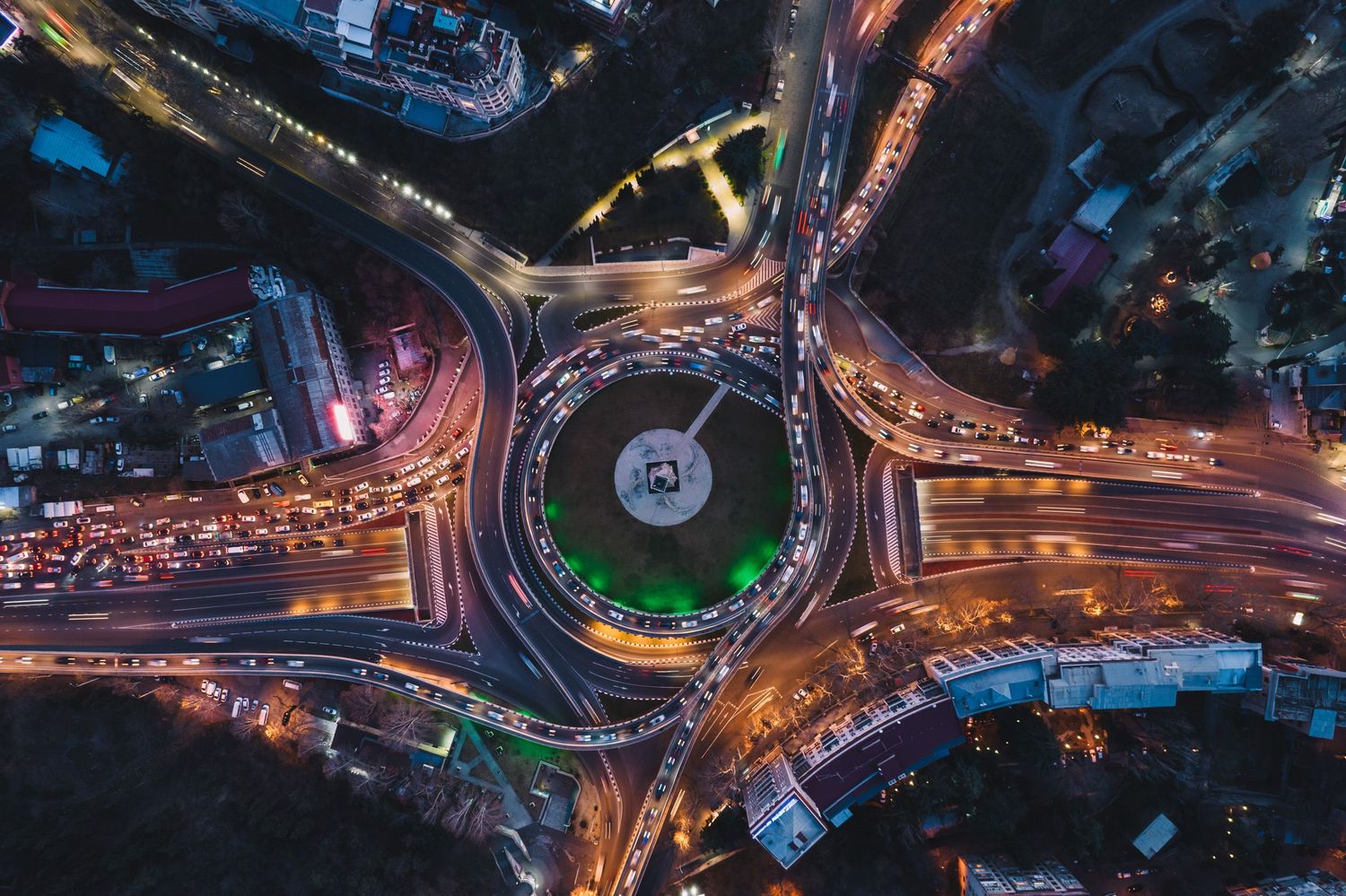 Night drone shot of a circular road junction lit with vibrant streetlights and moving traffic.
