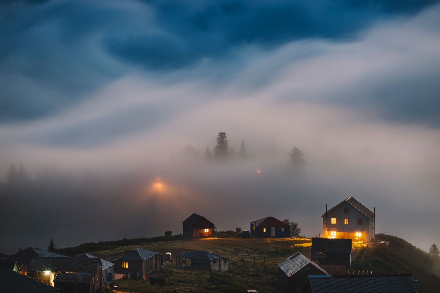 Mountain village at twilight covered in low-lying fog — glowing rooftops under a dramatic sky.