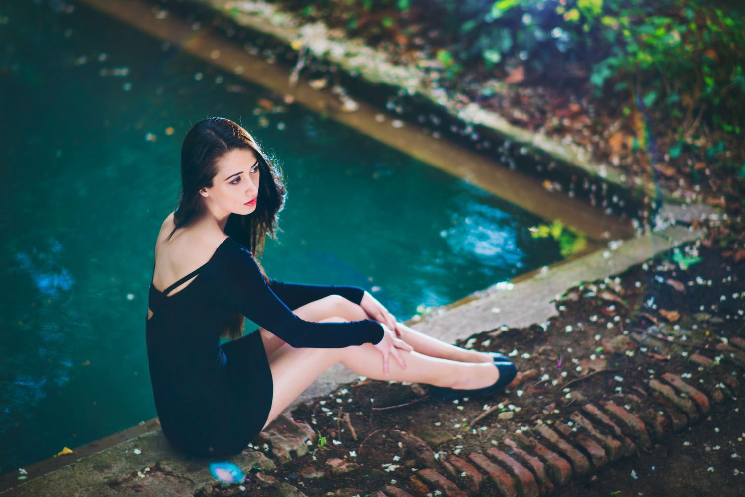 Young woman in black dress sitting by a turquoise pool — relaxed editorial-style portrait.