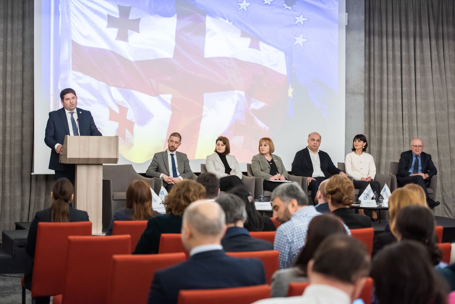 Conference or seminar in progress, with a panel of speakers and a large projected screen in the background.