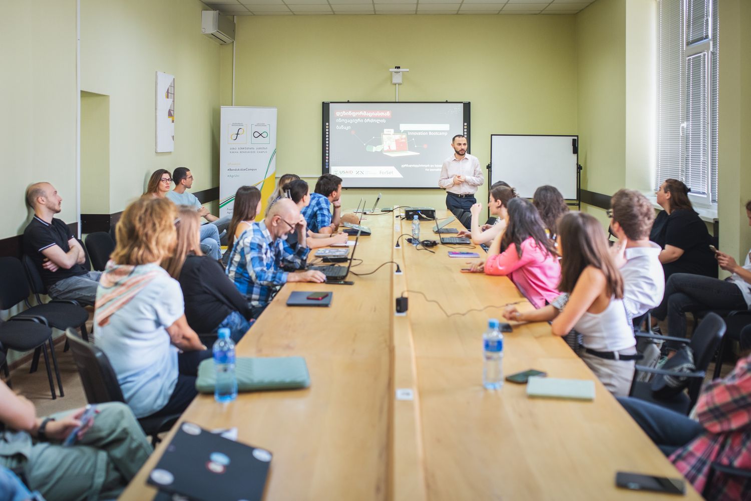 Large-scale training or seminar with participants seated along a long meeting table.