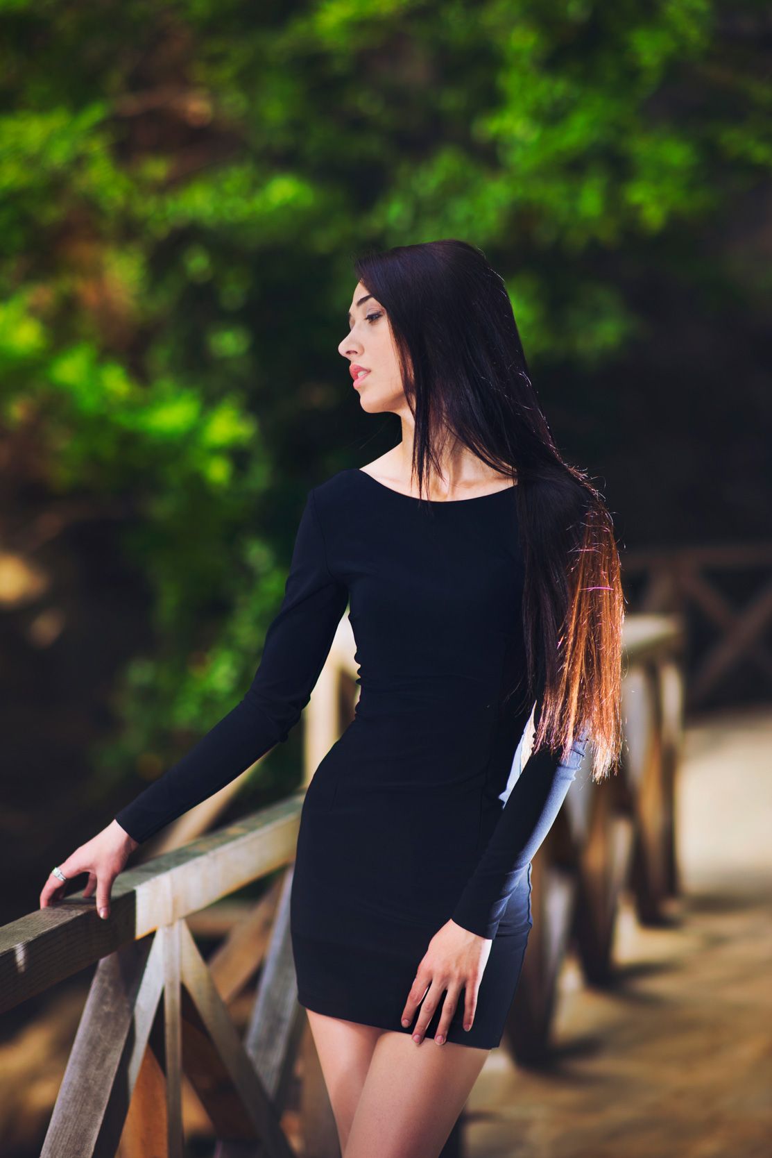 Young woman in a black dress standing beside a wooden fence — natural light portrait in greenery.