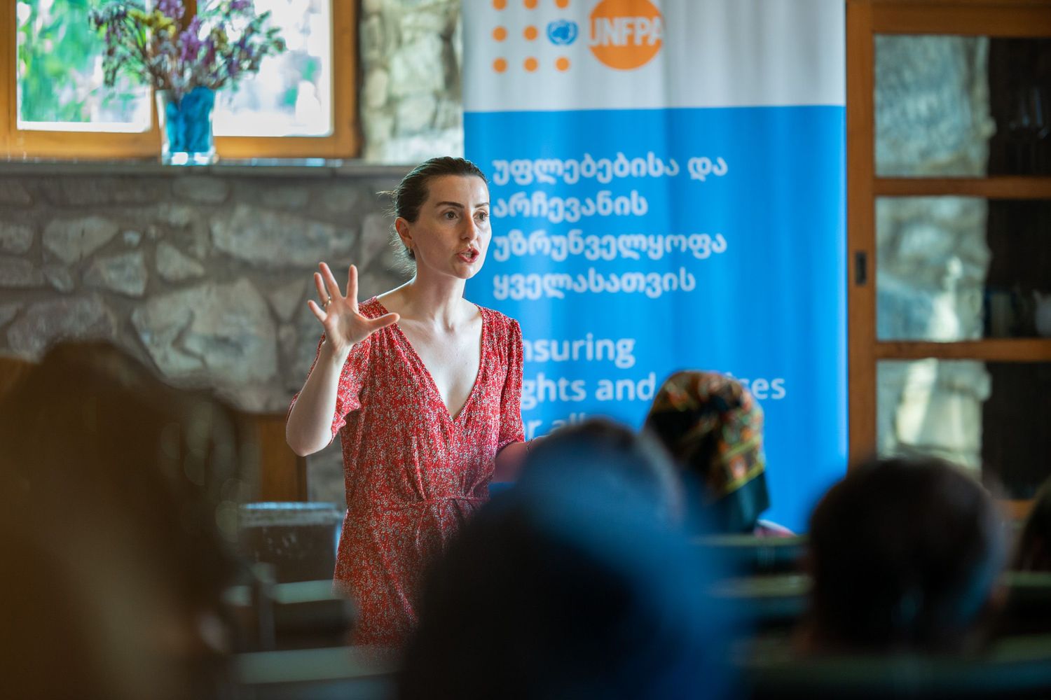 Speaker in a red dress addressing an audience during a formal indoor presentation.