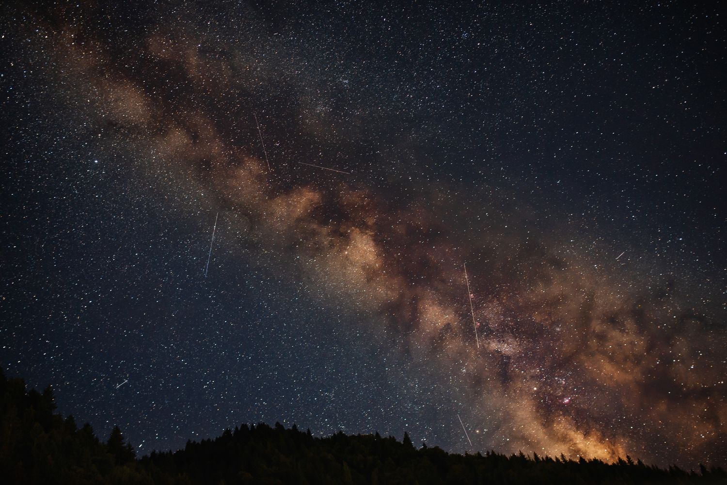 Milky Way stretches across the night sky above a silhouetted forested ridge.