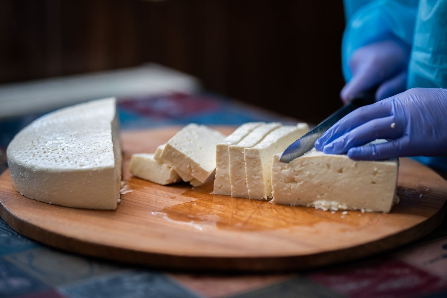 Chef slicing fresh traditional cheese on a wooden board with gloves.
