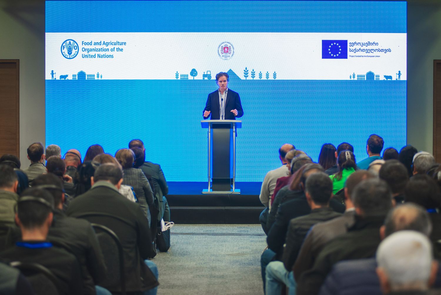 Formal conference setting with speaker at podium addressing a seated audience; EU and UN branding visible.