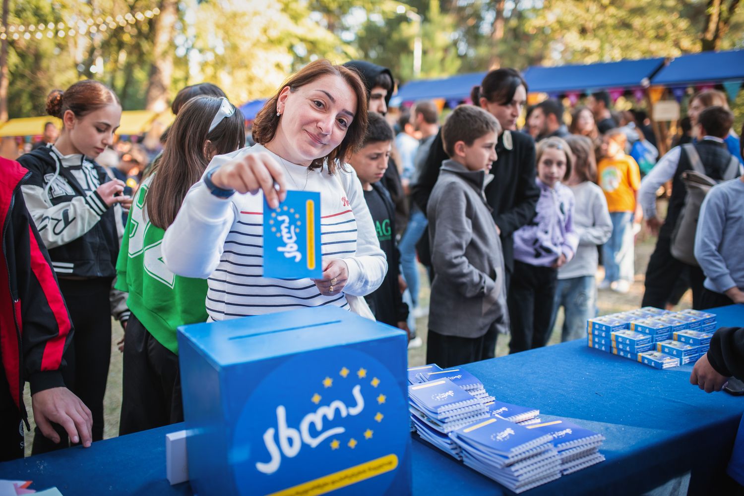 Young man smiling and presenting a product or leaflet at a public outdoor festival booth.