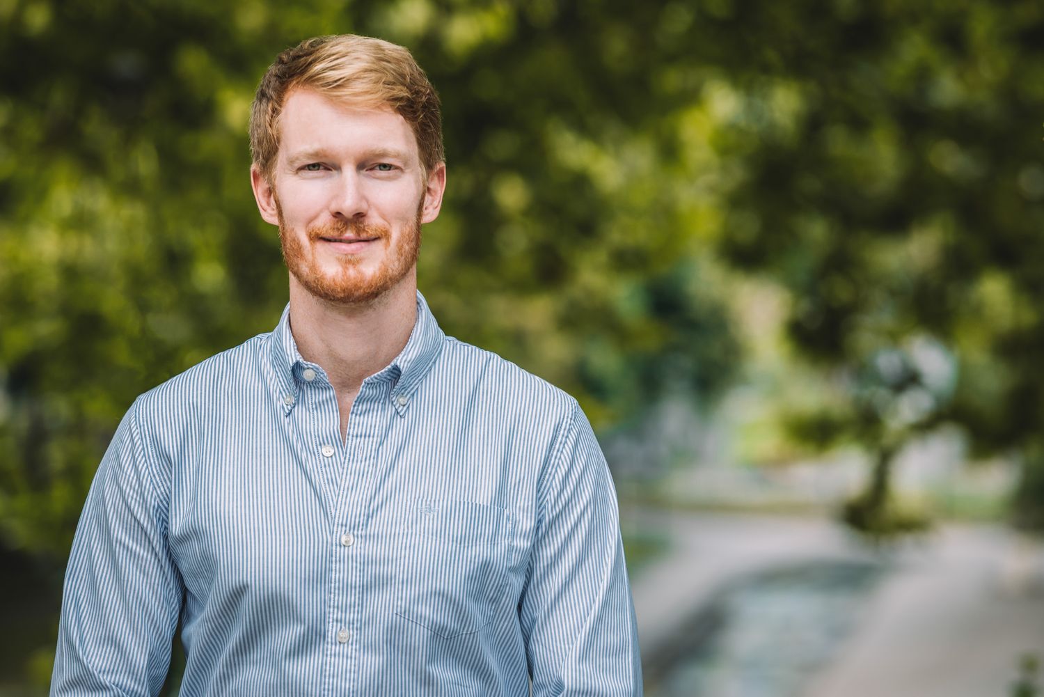 Portrait of a young man smiling confidently outdoors with greenery in the background.