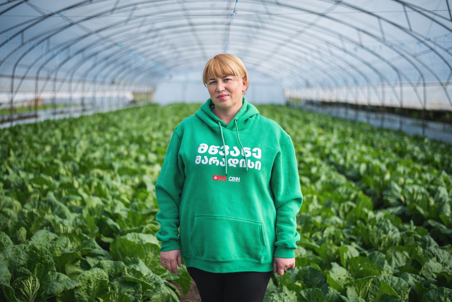 Young person standing inside a greenhouse surrounded by rows of leafy greens.