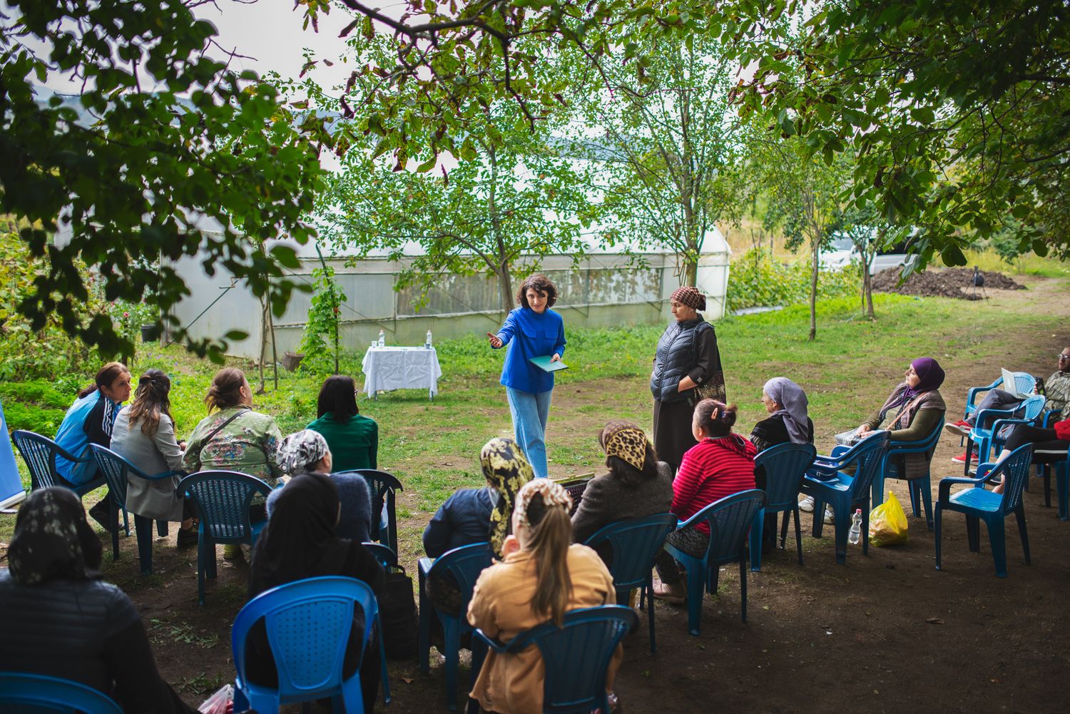 Outdoor community gathering in a park setting, with a person addressing seated attendees.