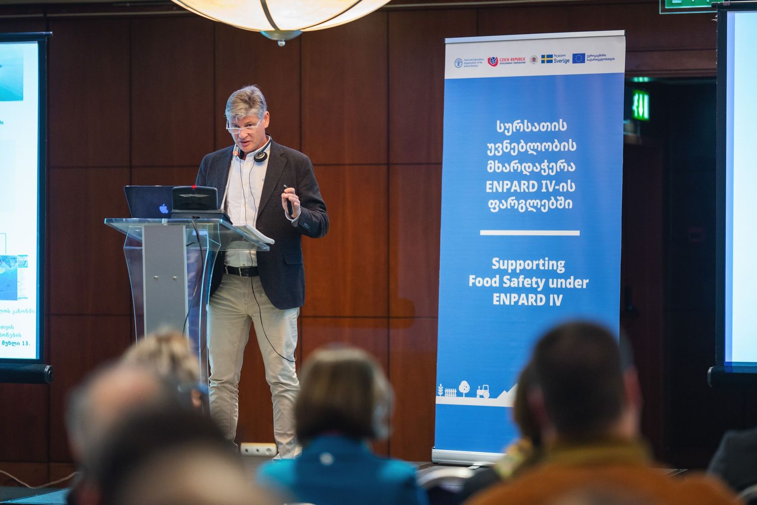 Professional speaker delivering a presentation with a blue digital screen in the background.