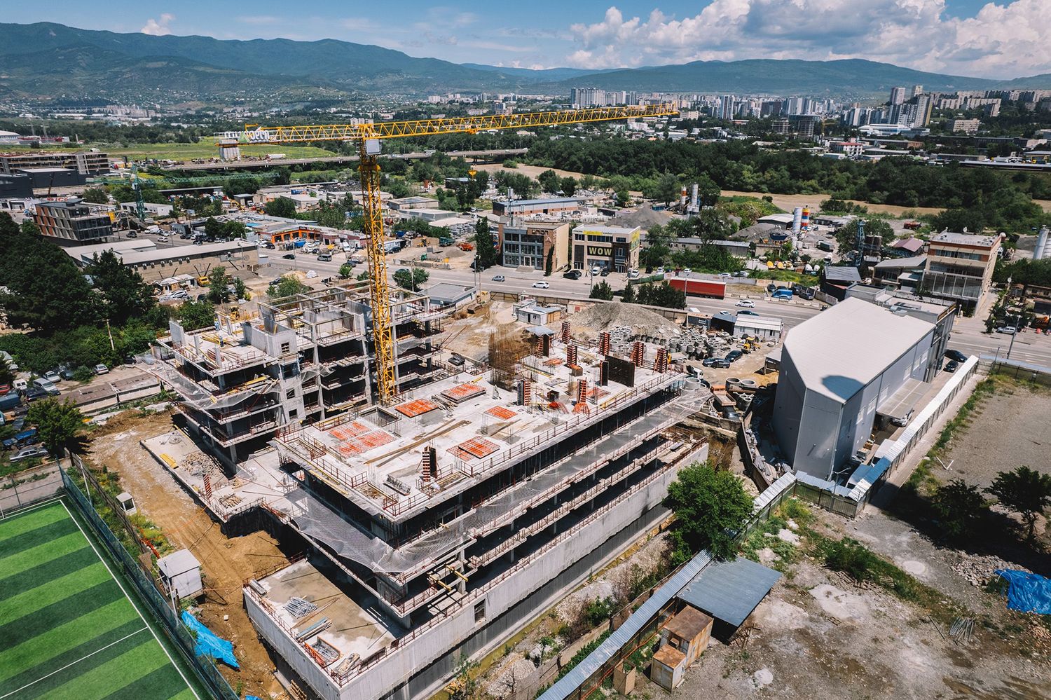 Aerial view of an industrial construction site with cranes, buildings, and a distant city skyline.