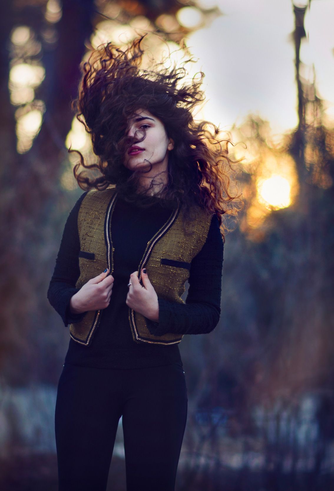 Moody portrait of a woman in black coat standing in a sunlit forest path.