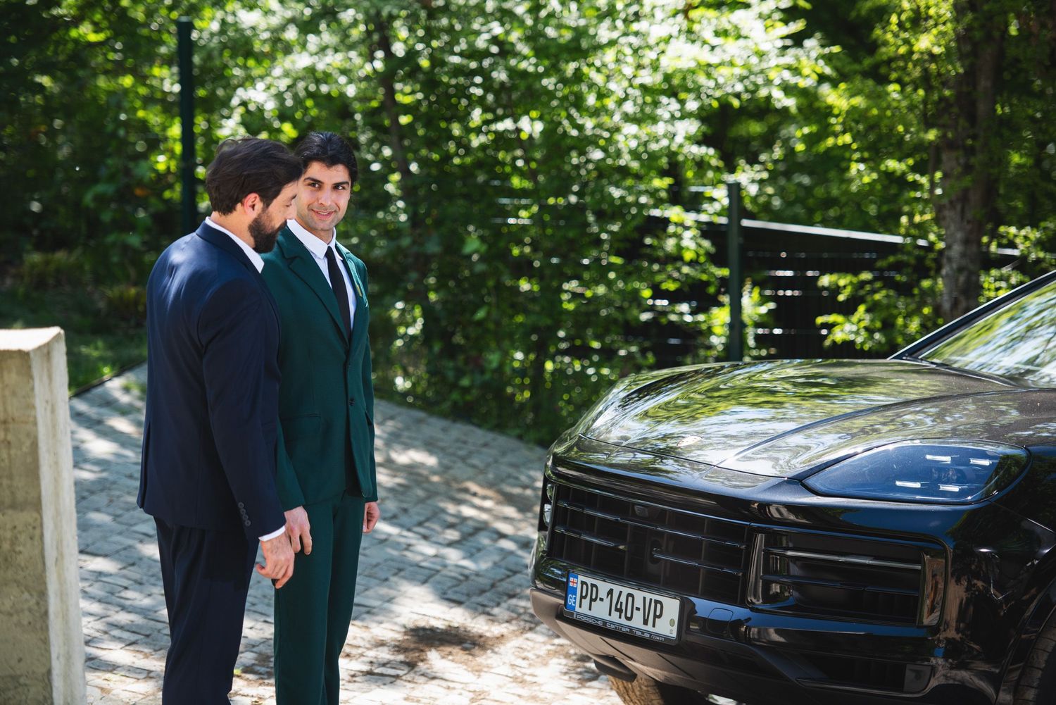 Businessman in green suit talking with a man near a black SUV — casual conversation moment.