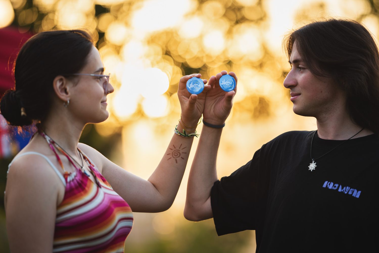 Two young women smiling and clinking dumbbells outdoors during a sunny workout session.