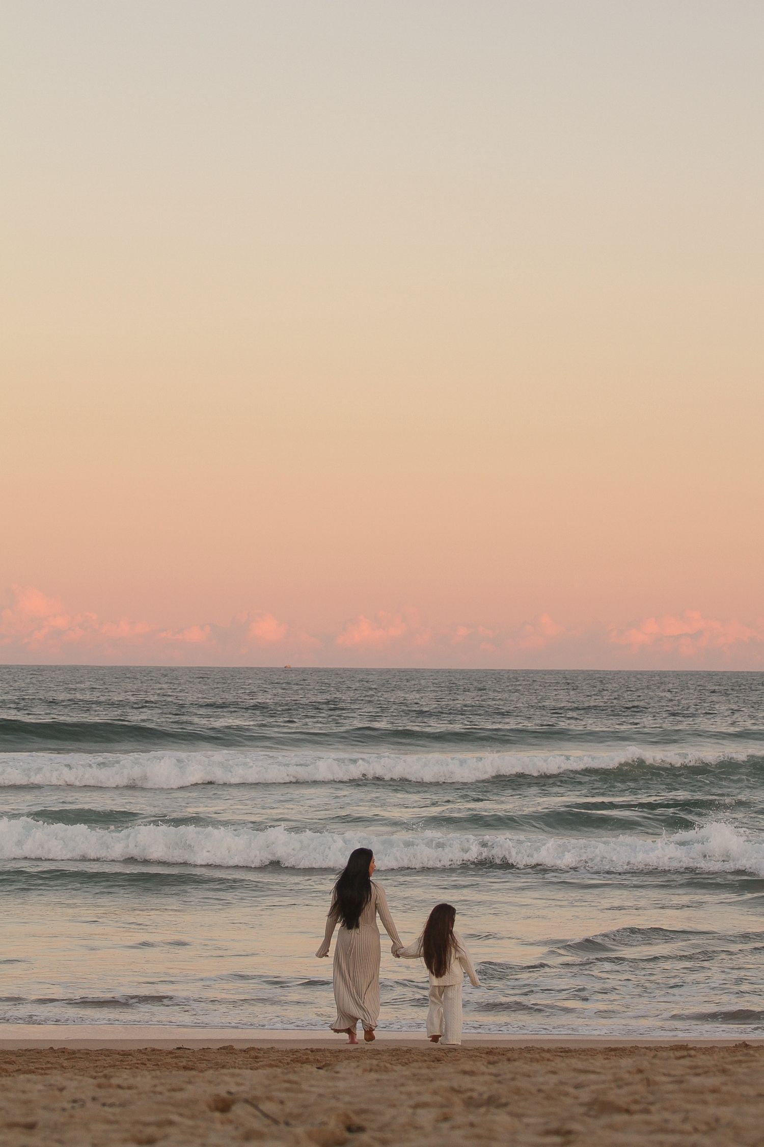 Two people in white outfits walk along a peaceful beach at sunset with pink clouds.