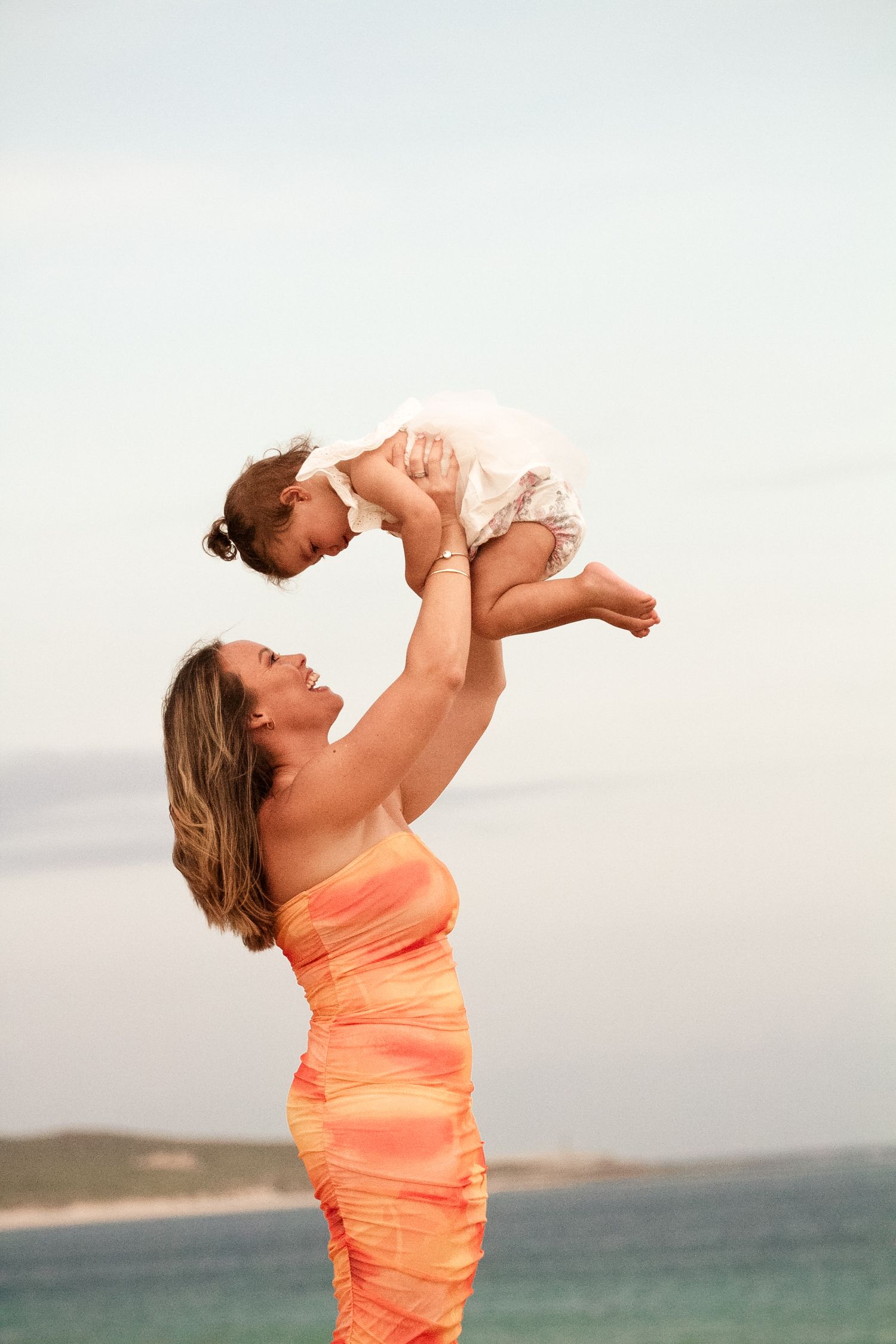 Parent lifts baby high into the air while wearing a bright coral dress against an ocean backdrop.