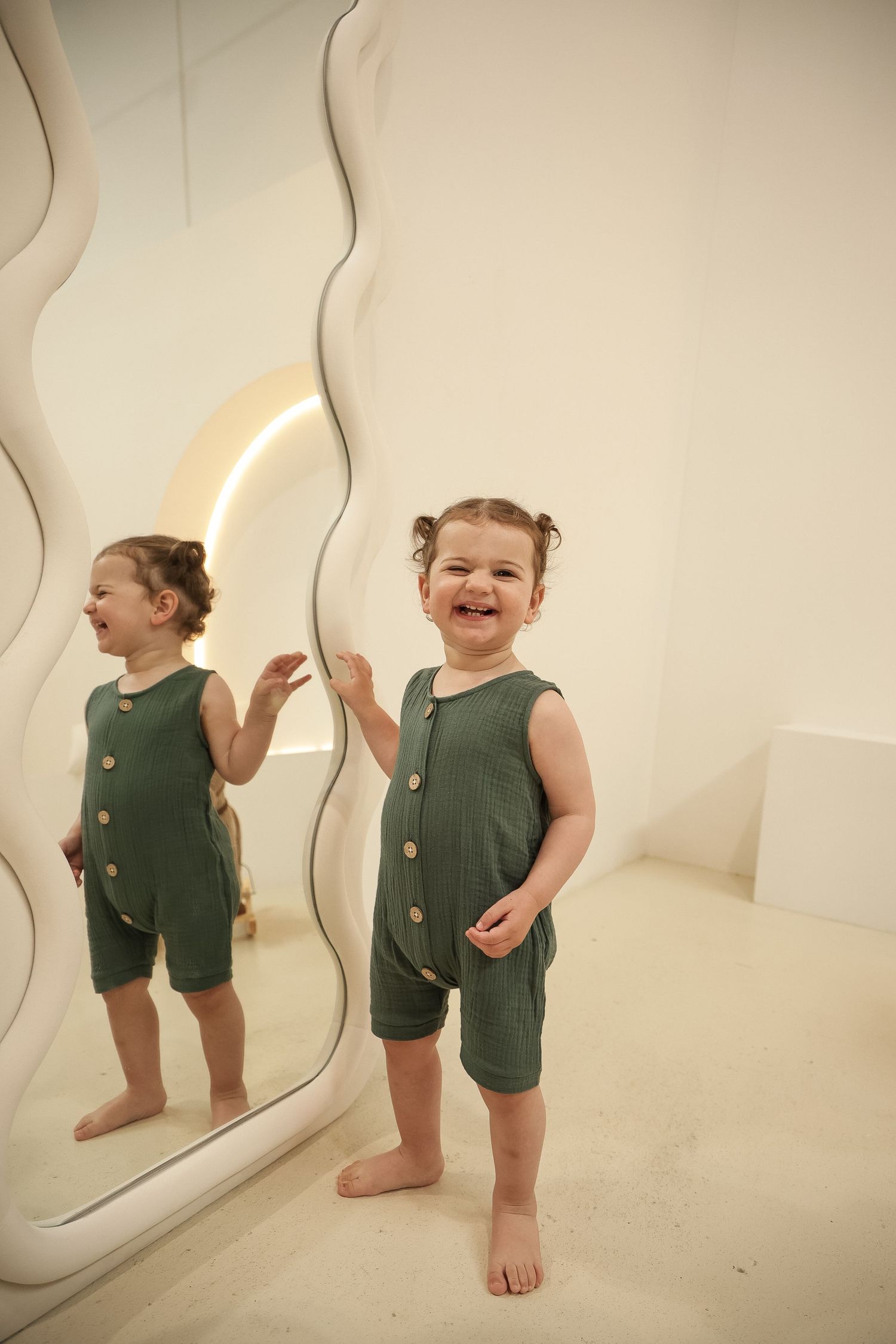 Young child in olive green romper playfully stands next to a wavy mirror in a minimalist white room.