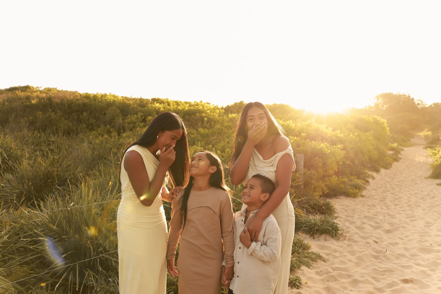 A group laughs together on a sandy path during golden hour sunset with sunlit grass in the background.