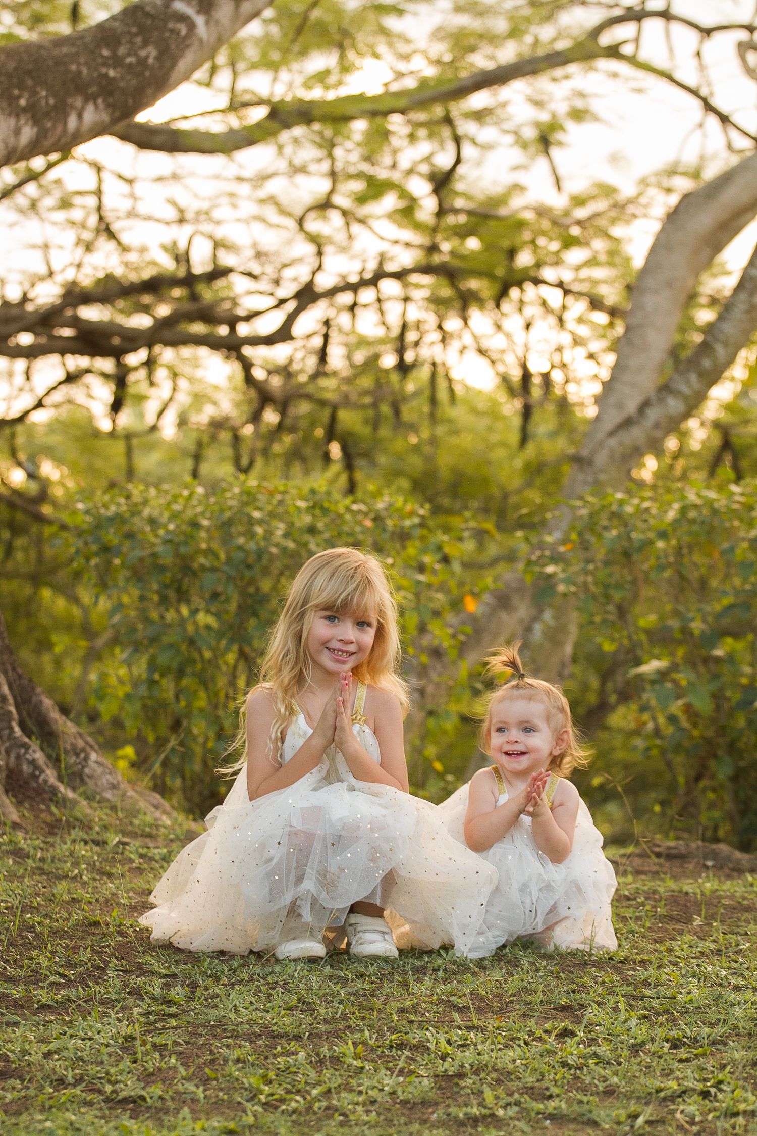 Two girls in white dresses pose playfully on grass under trees at sunset.