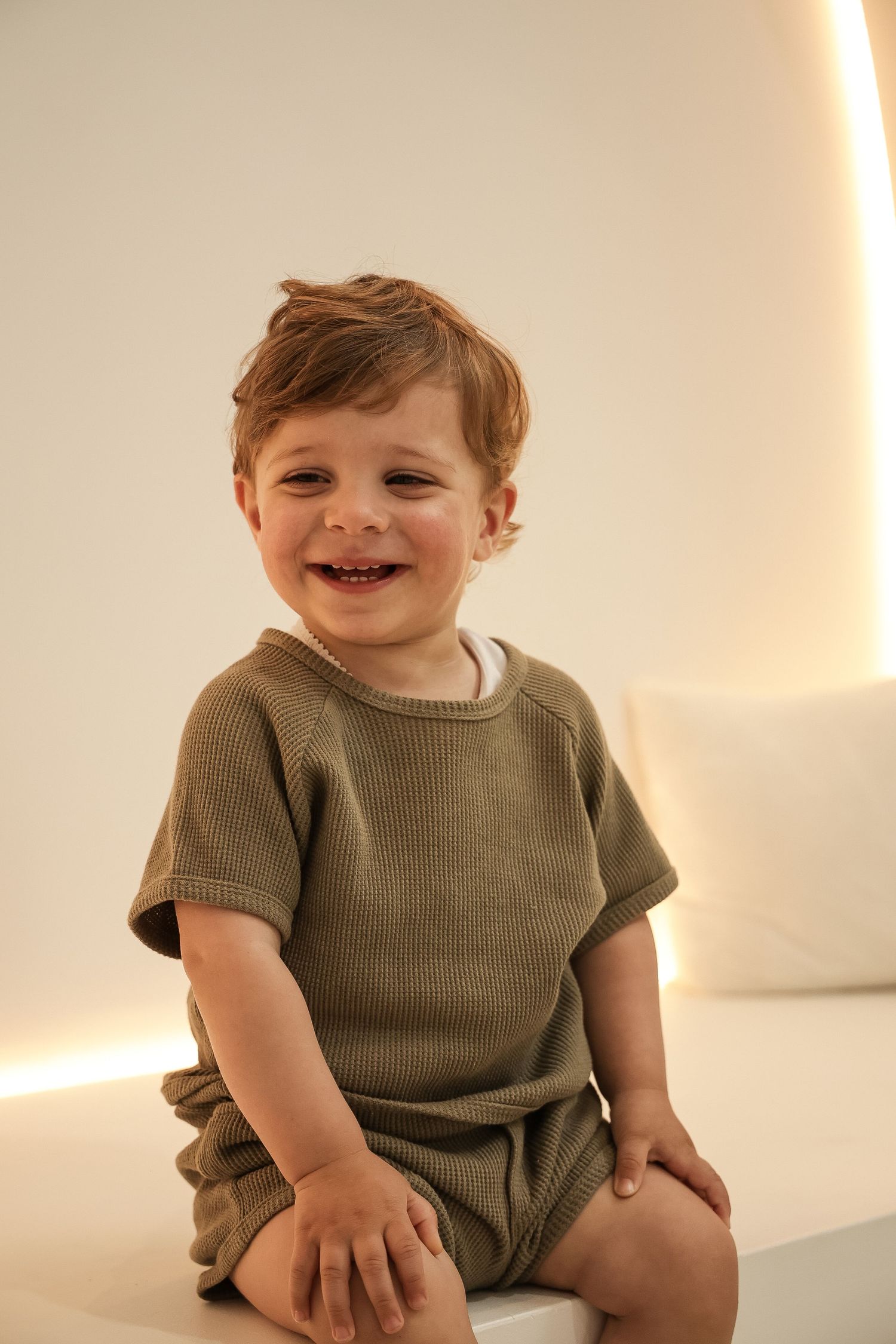 Small child wearing a neutral-colored ribbed romper sits smiling in a softly lit room.