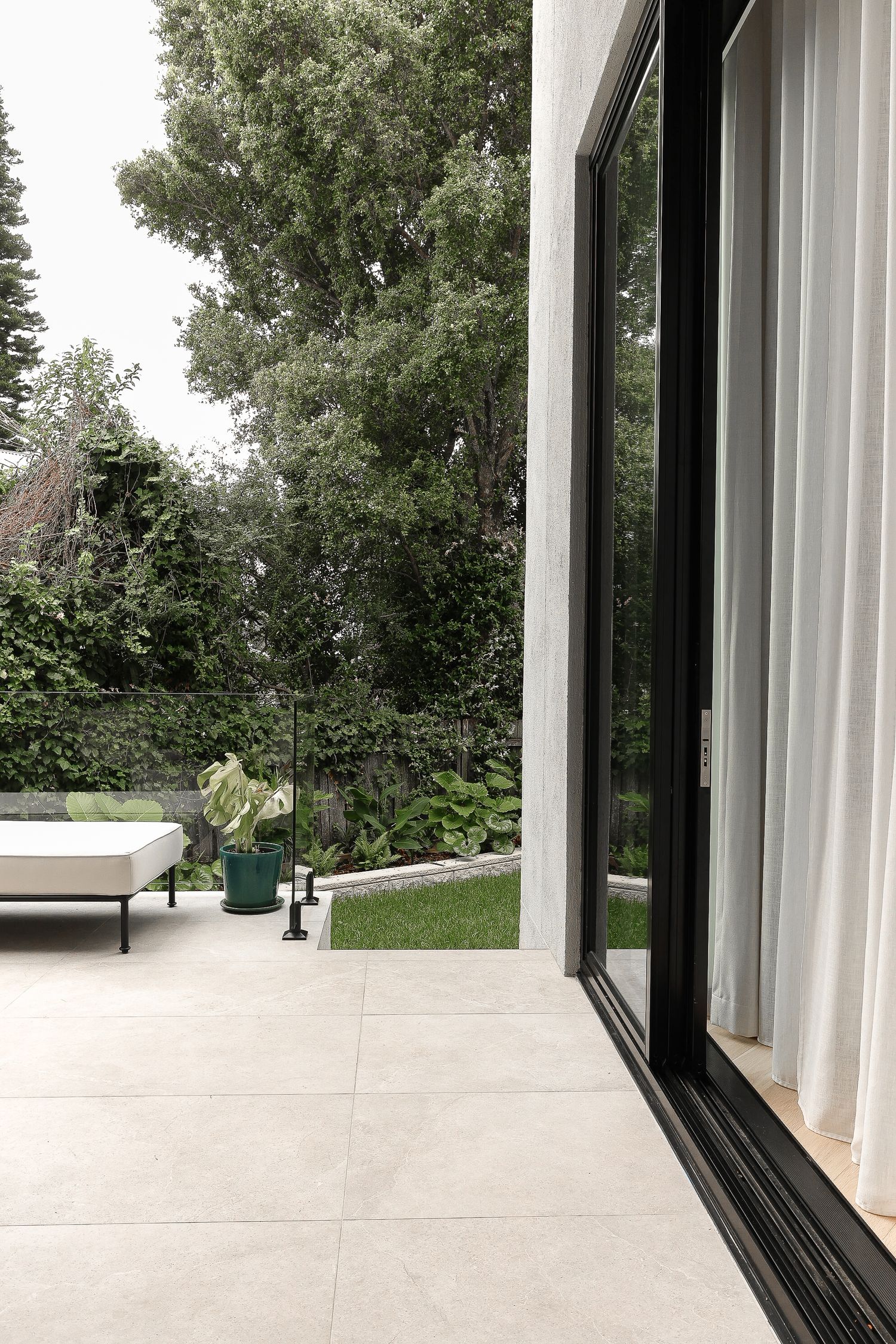 Modern white outdoor patio space with floor-to-ceiling windows overlooking lush garden landscape.