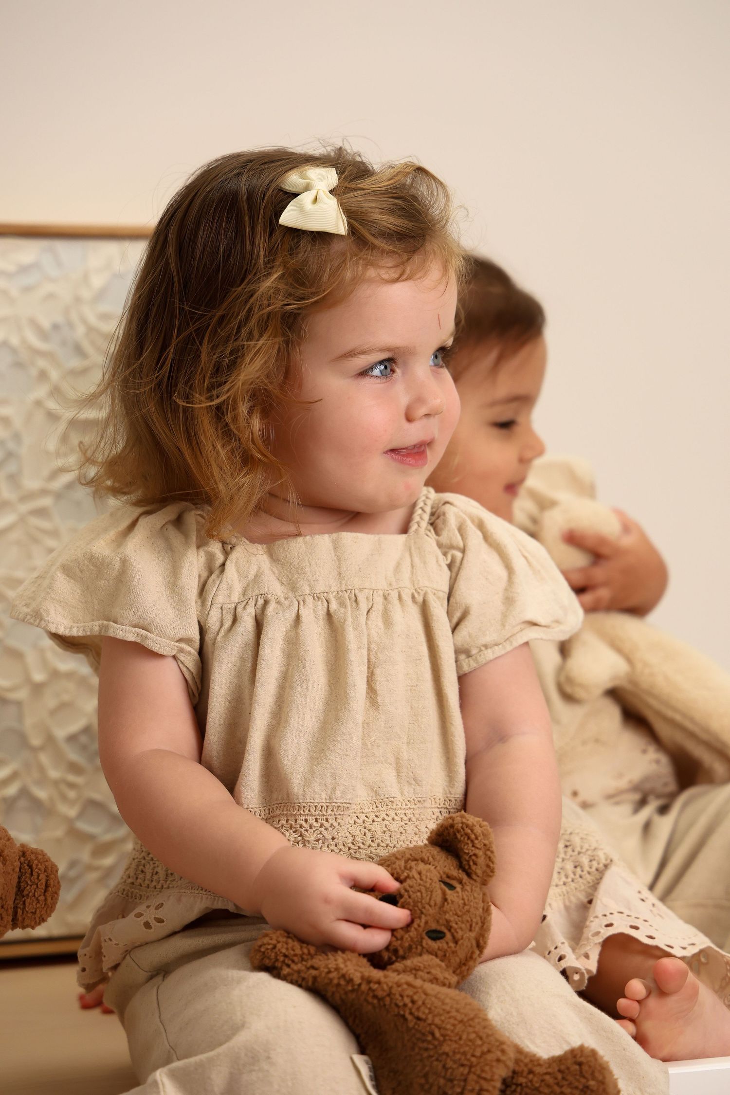 Young child in a beige dress holding a brown teddy bear while sitting on a patterned chair.