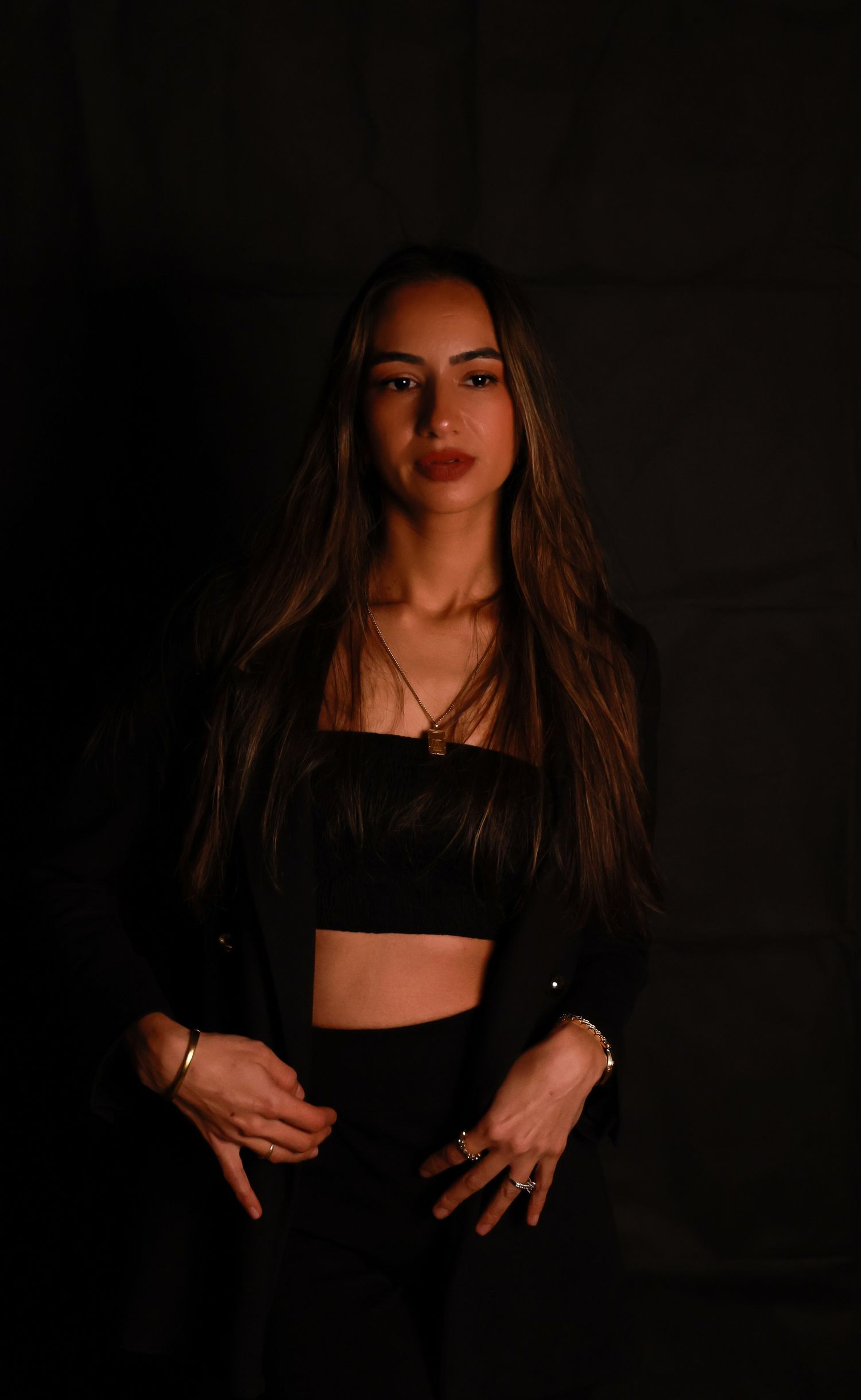 Low-key portrait of woman in black crop top and jacket with long hair against dark wall.