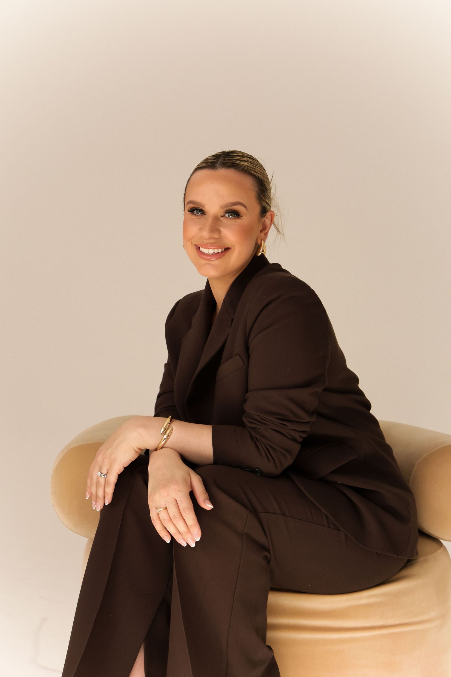 Person in dark blazer posing on beige ottoman against white background.
