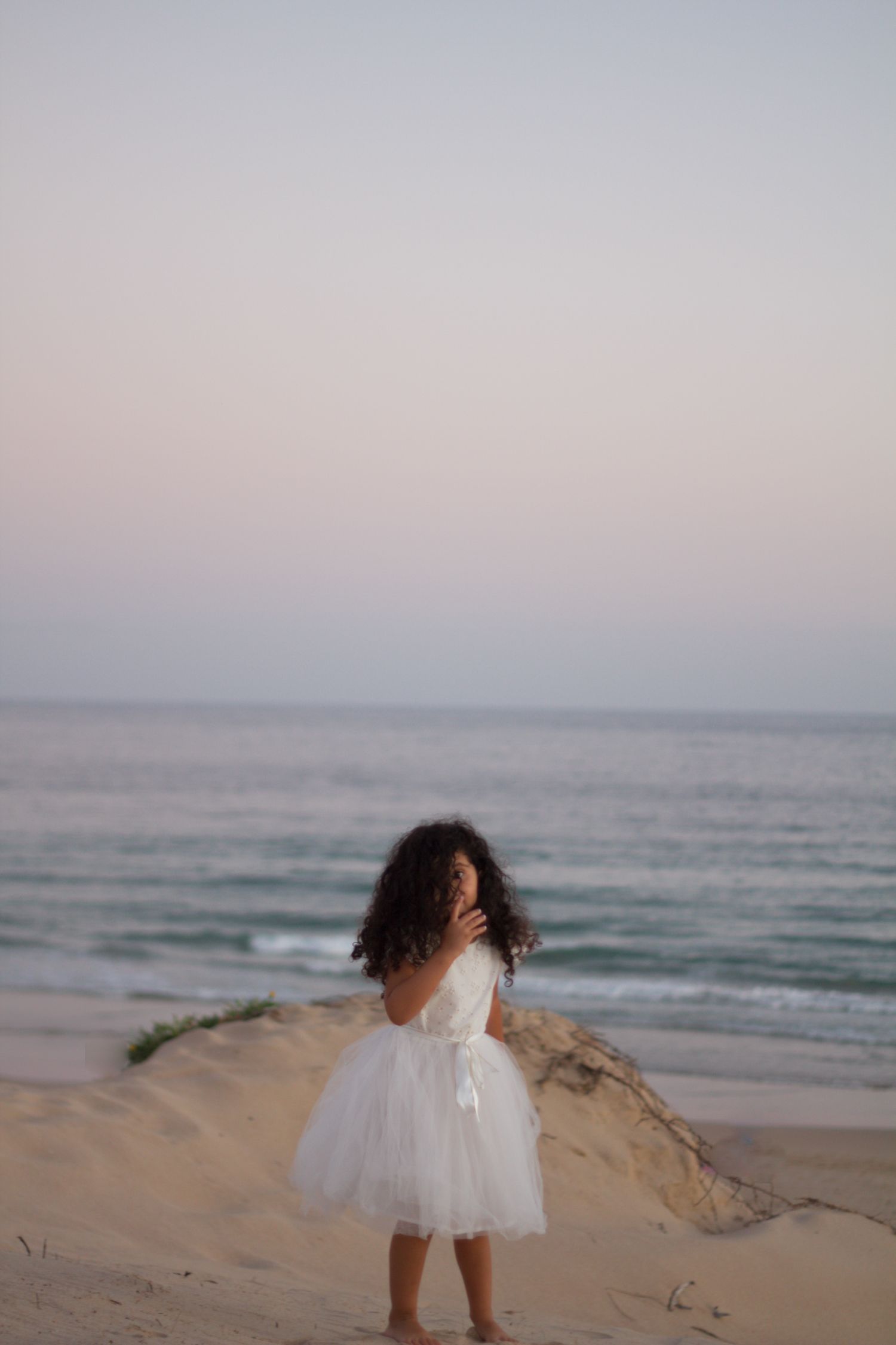 A child in a white dress twirls playfully on a sandy beach at sunset with ocean waves in the background.