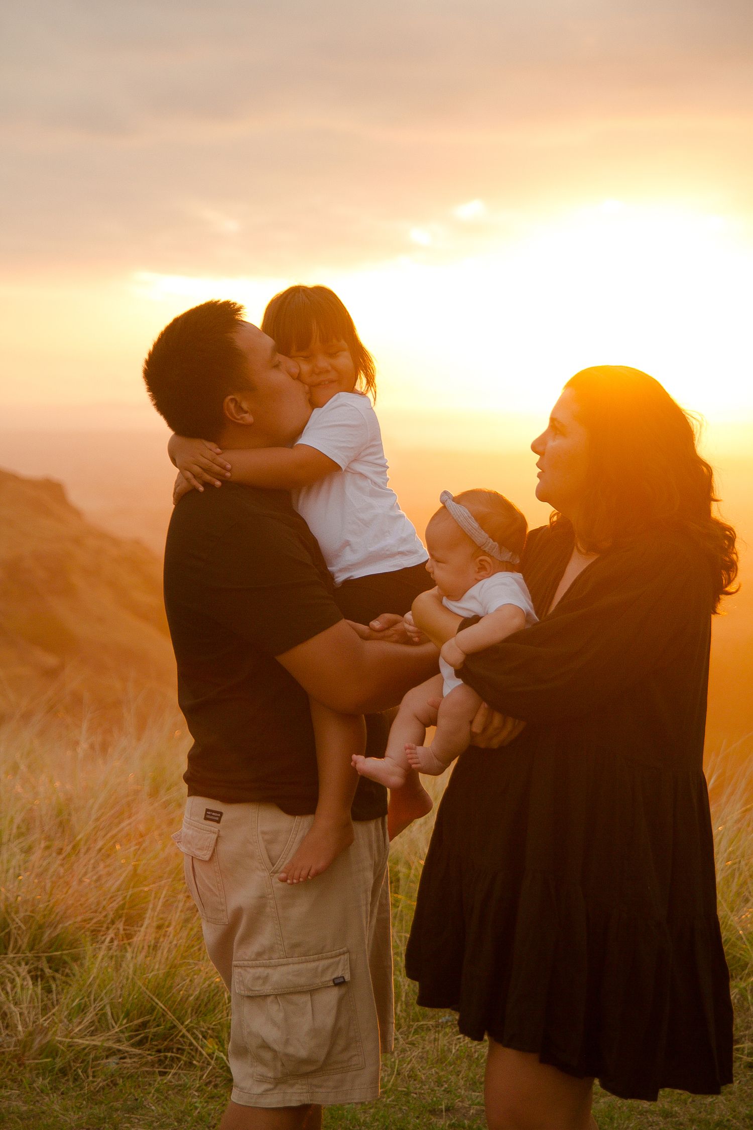 Family shares tender moment at sunset on grassy hilltop.