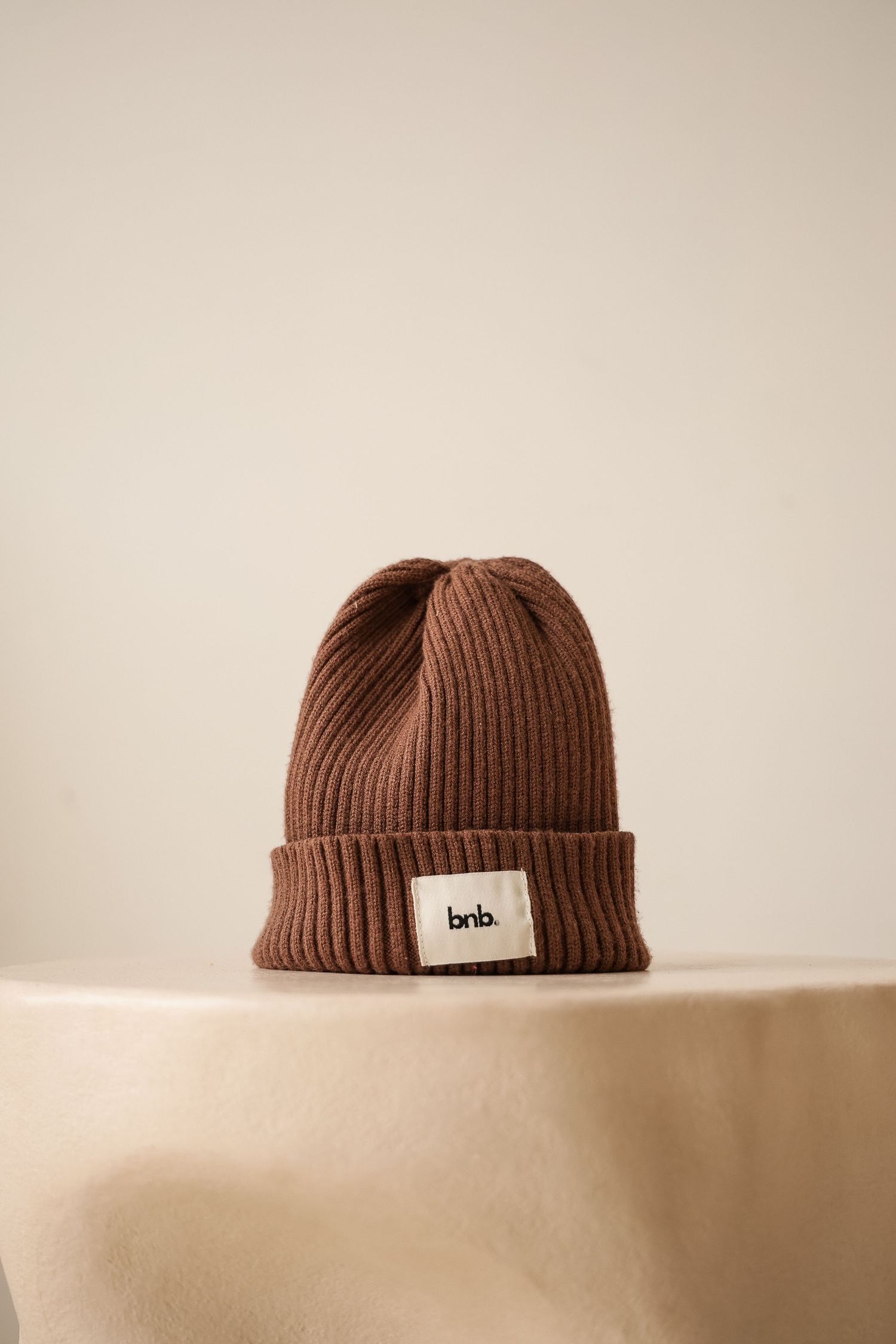 Brown ribbed beanie hat with bnb label patch displayed on beige surface.