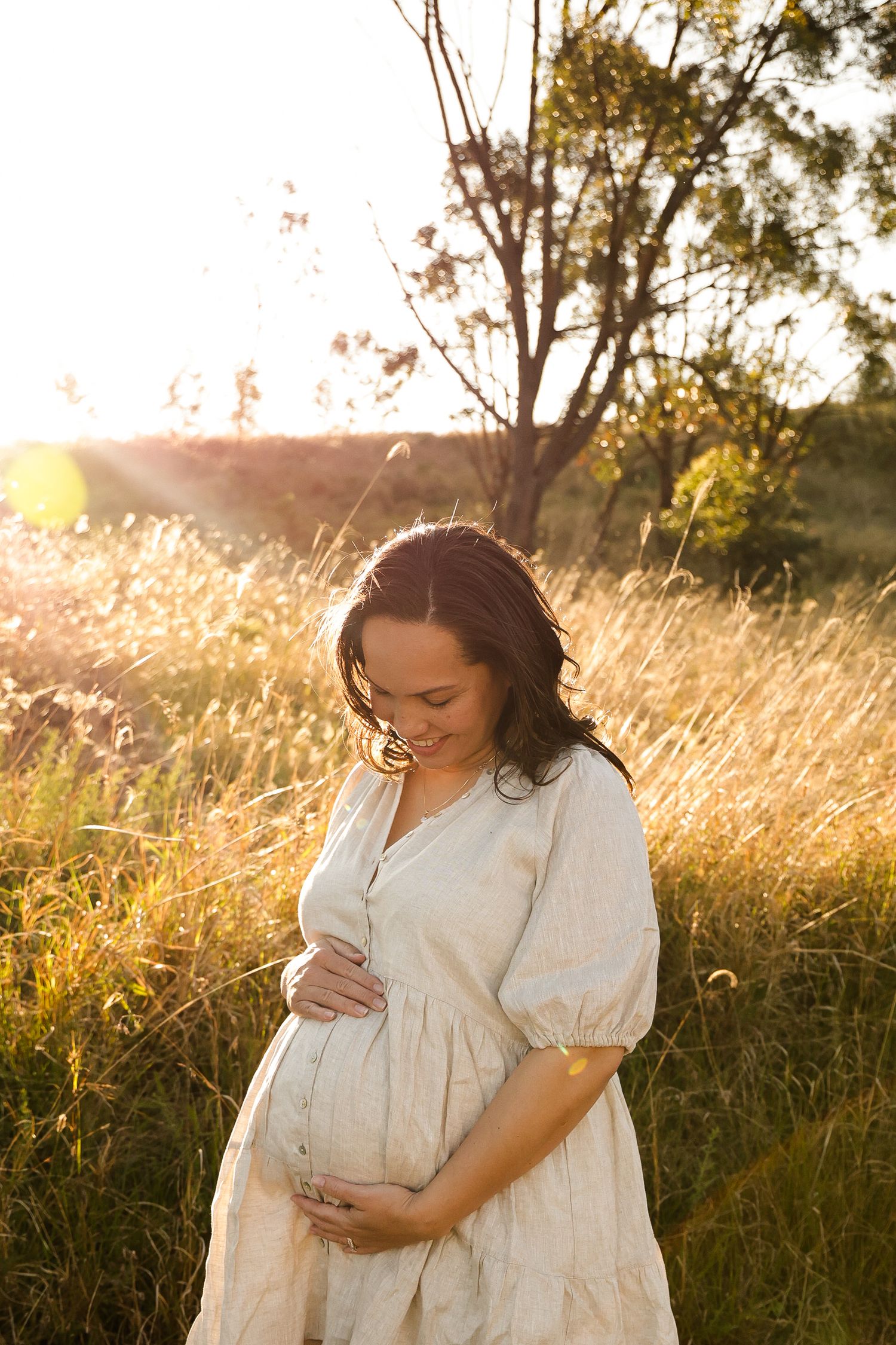 Expecting mother in white dress touches her belly during golden hour in tall grass field.
