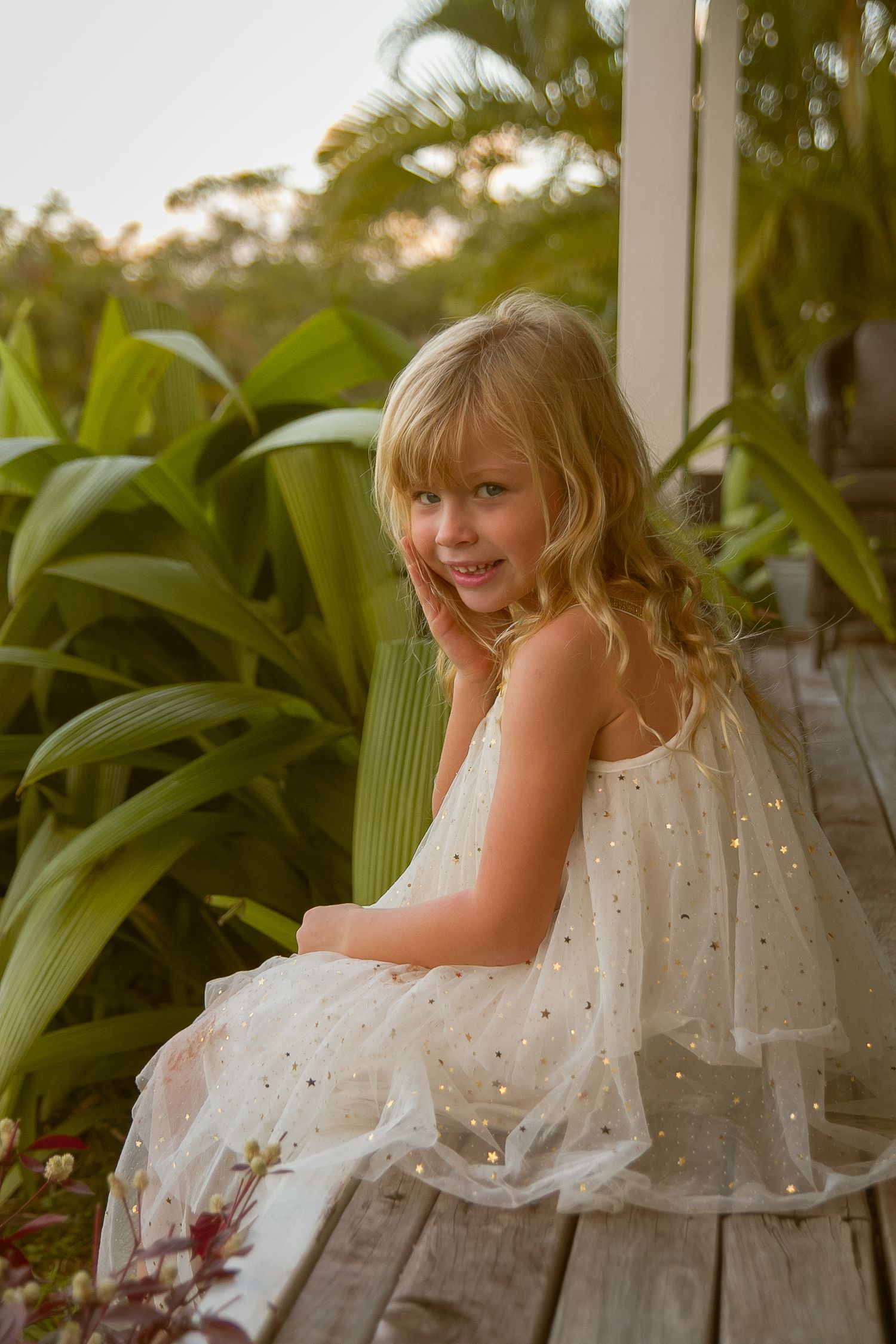 Young girl in white dress sits on wooden deck surrounded by tropical plants.