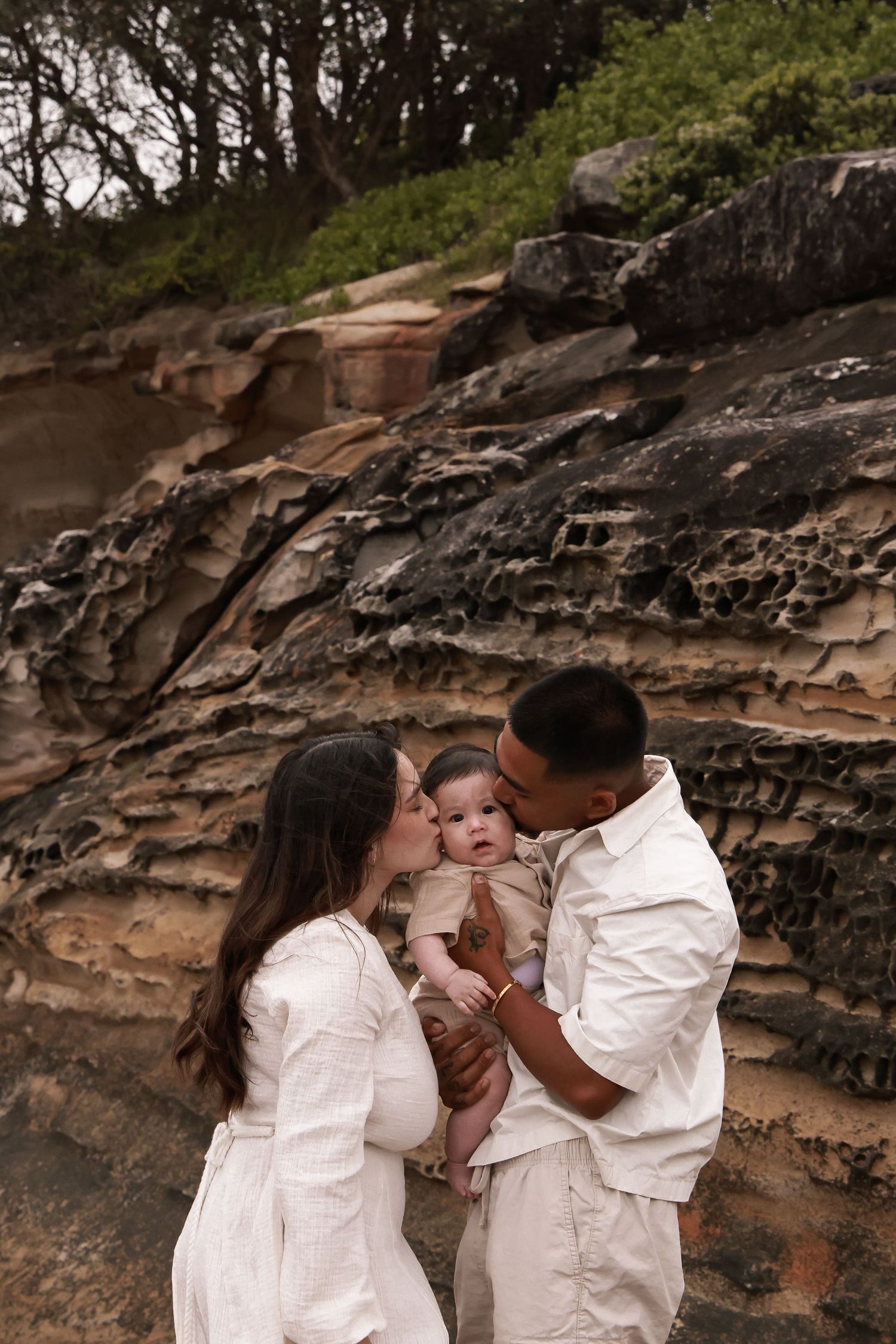 Parents in white outfits share a tender moment with their baby against layered rock formations.