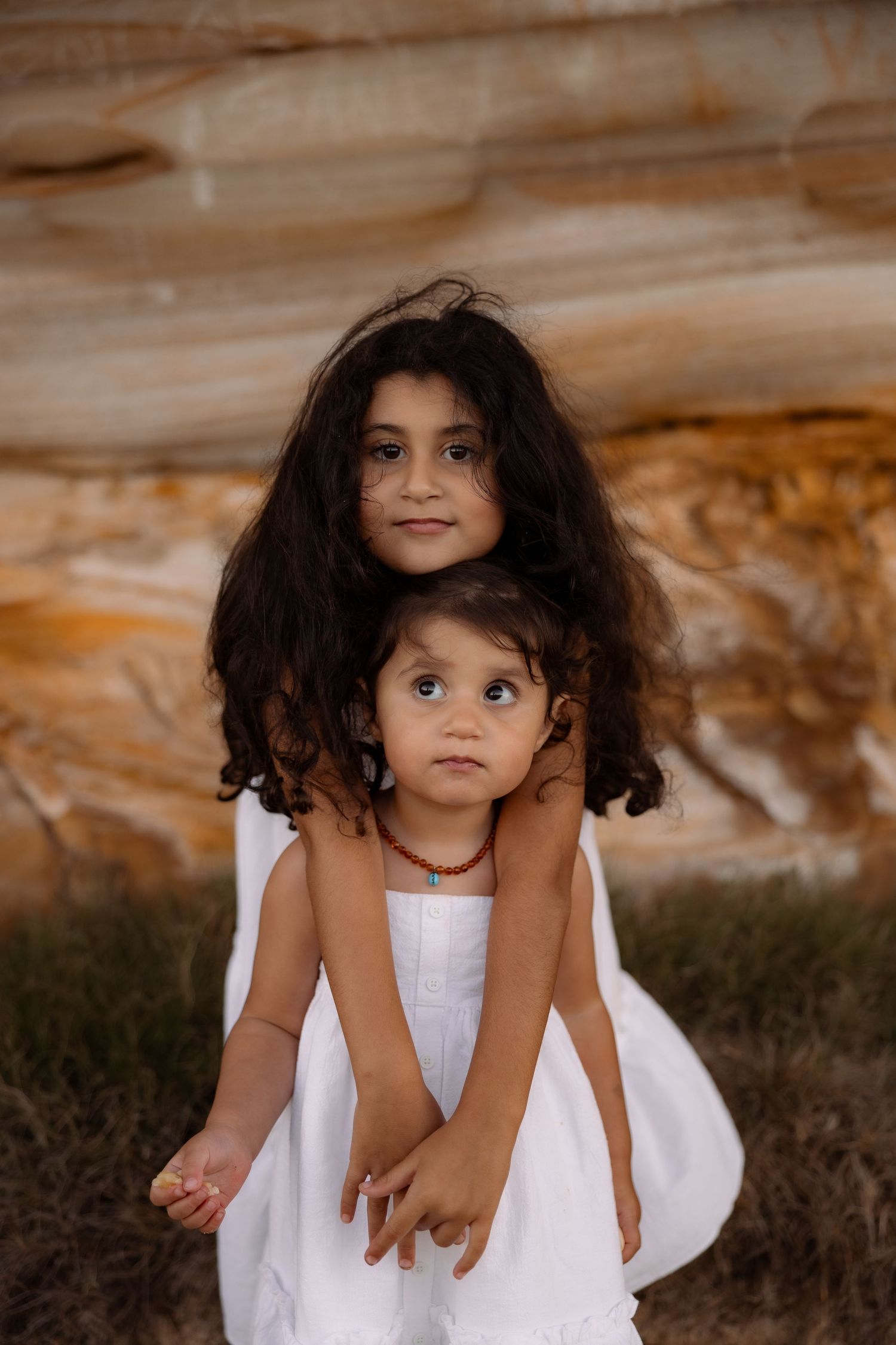 Two young girls in white dresses embrace outdoors near sandstone rocks, the older girl hugging the younger from behind.