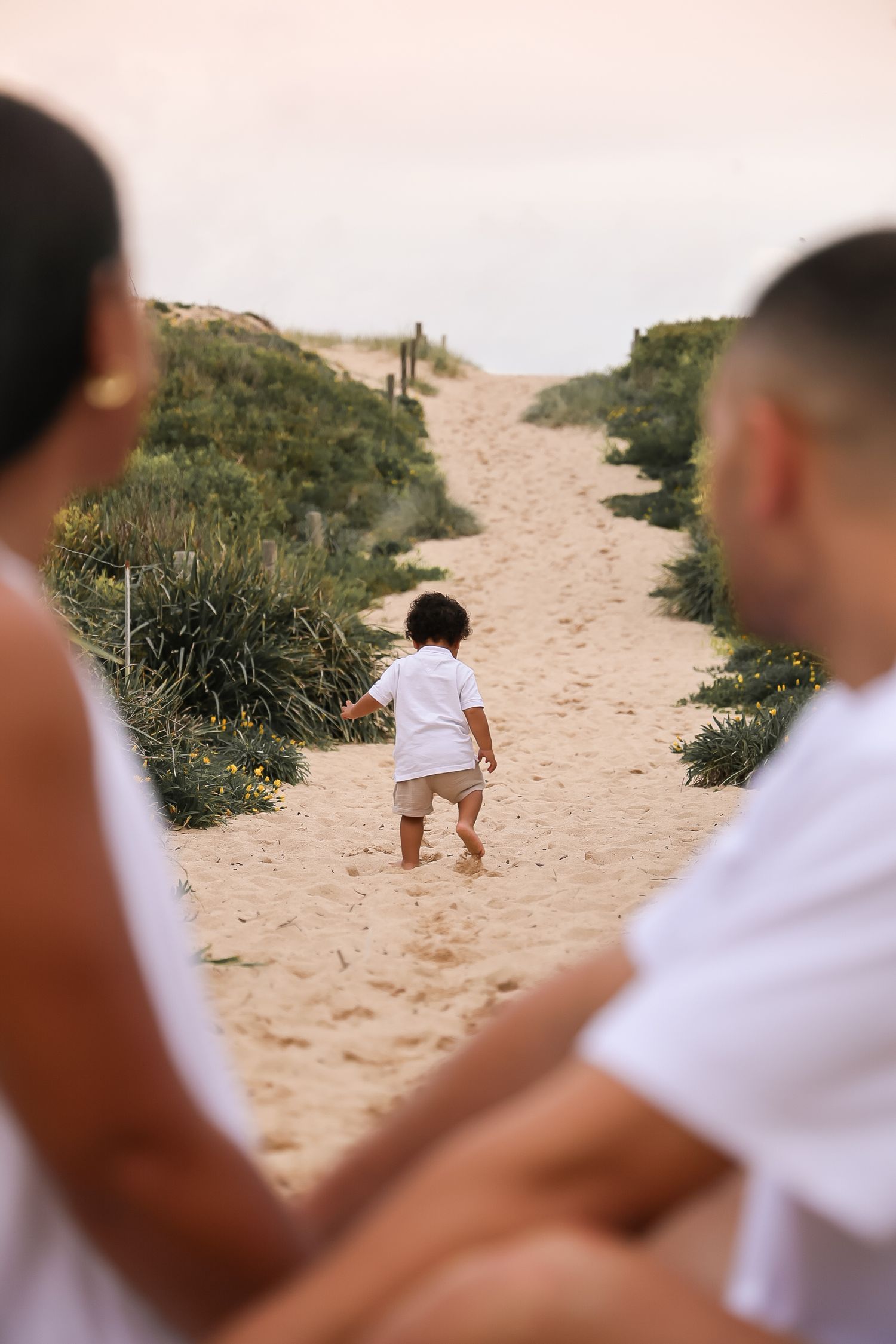 A toddler in white walks along a sandy beach path between coastal vegetation.