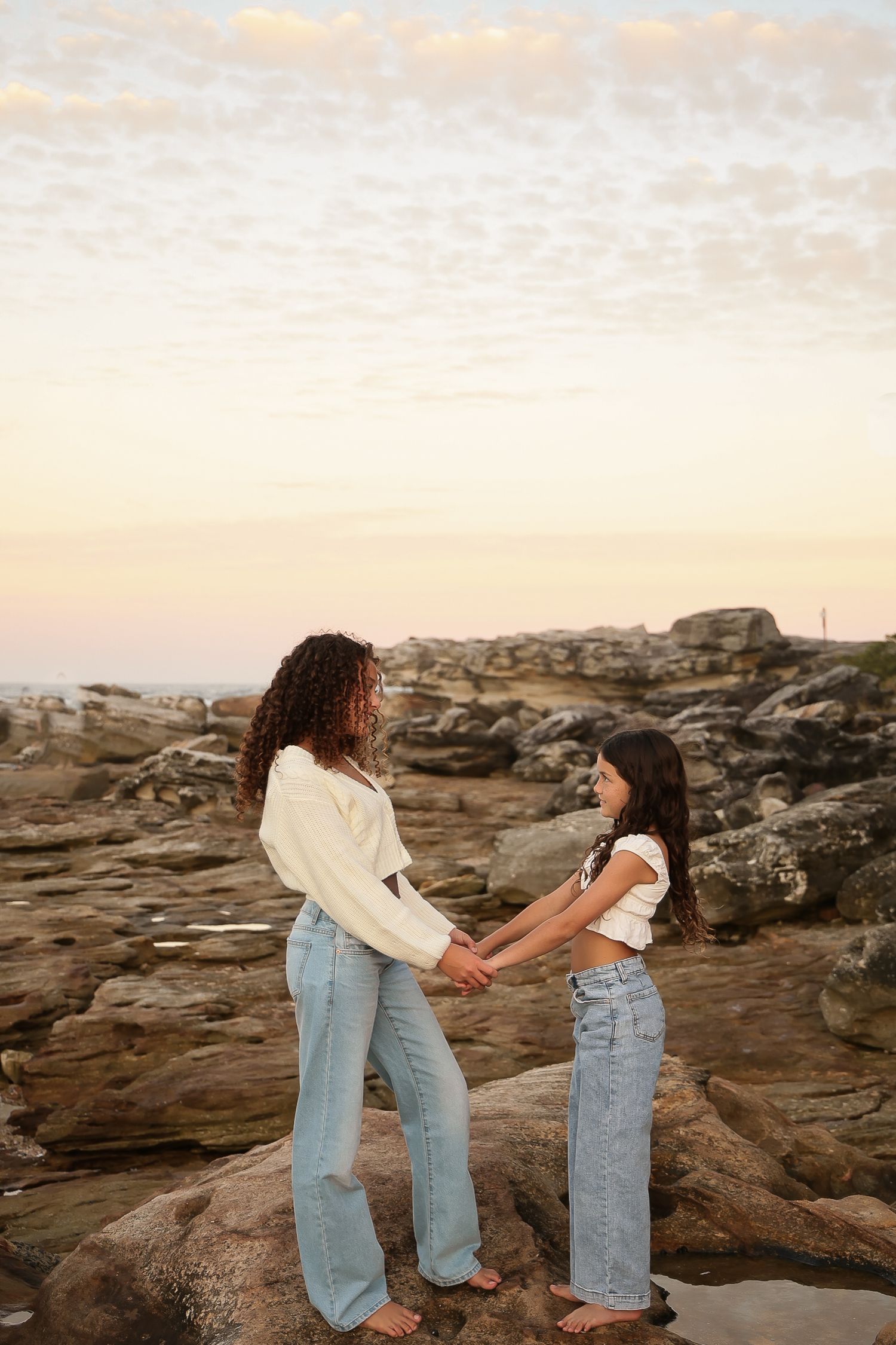 Two people holding hands while standing on rocky coastal formation at sunset.