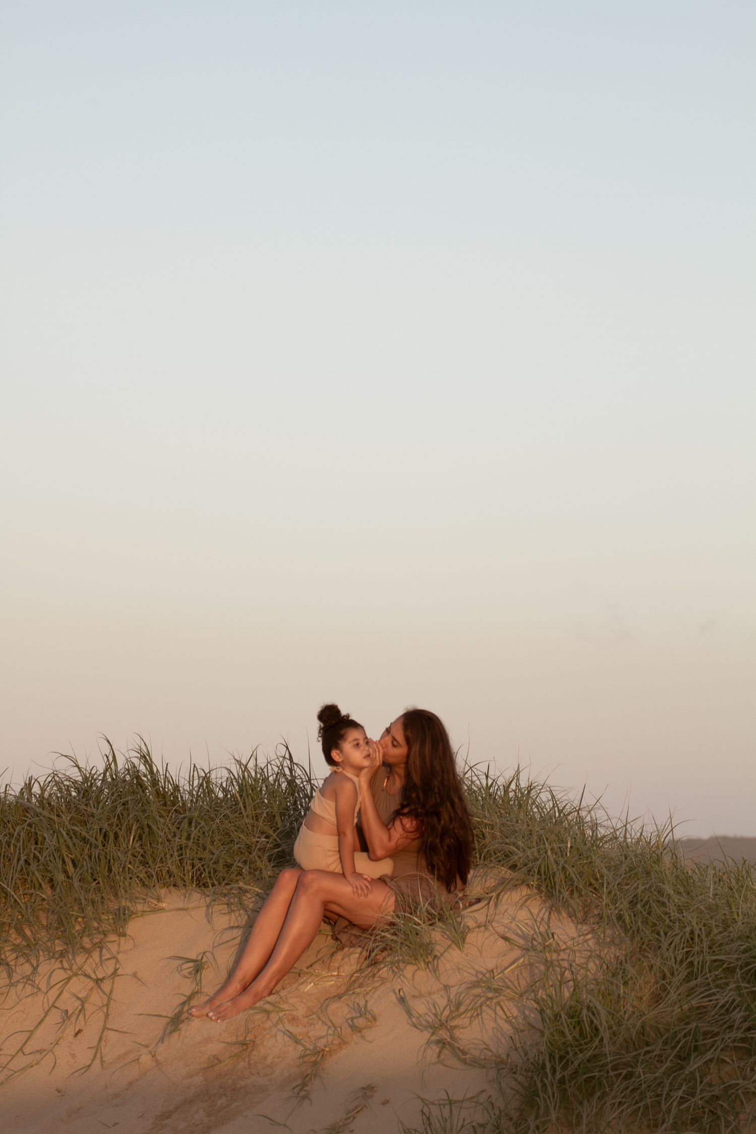 Mother and child share intimate moment on sandy dune at sunset.