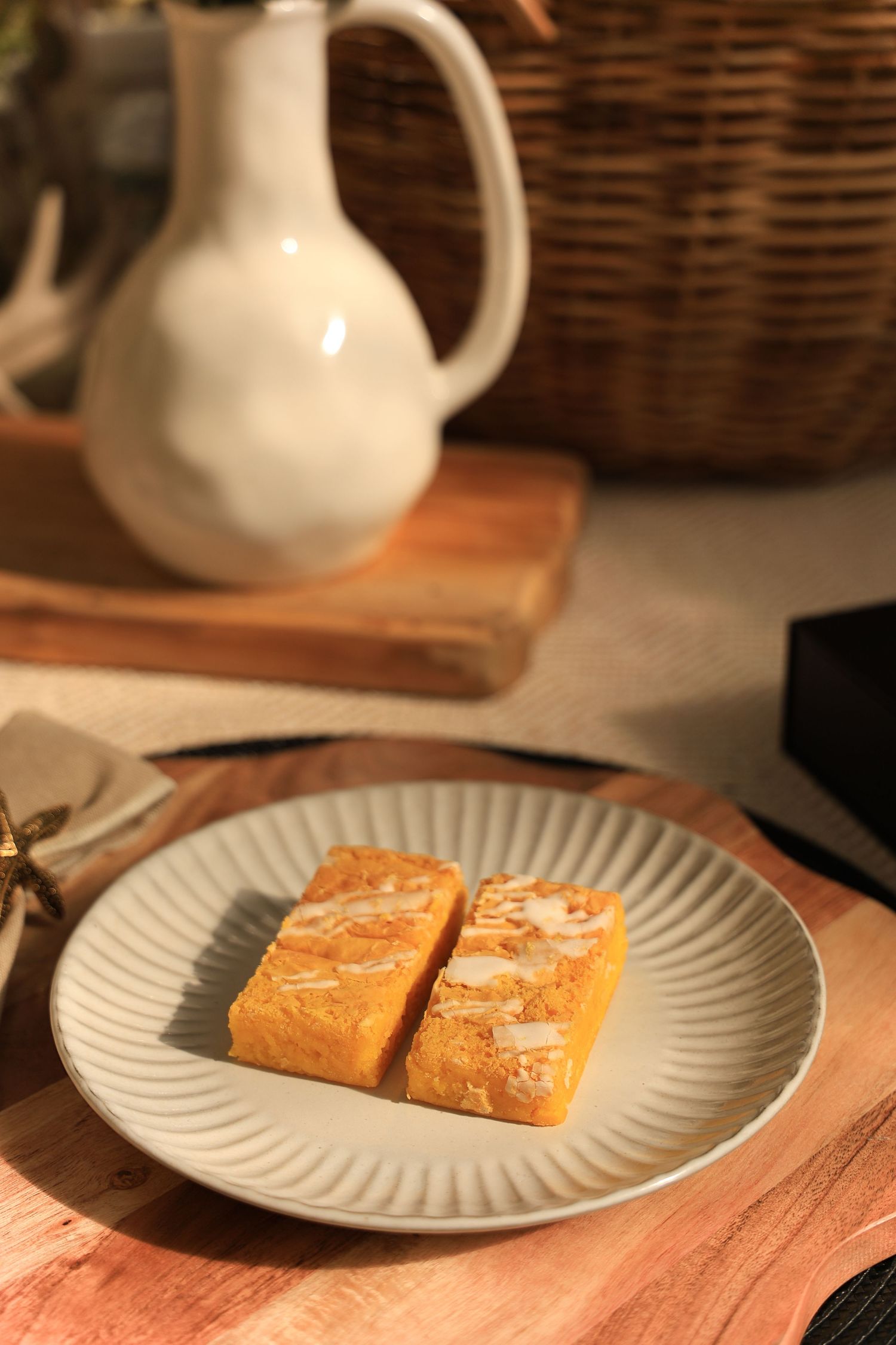 Two golden dessert bars on ribbed plate with white ceramic pitcher in background.